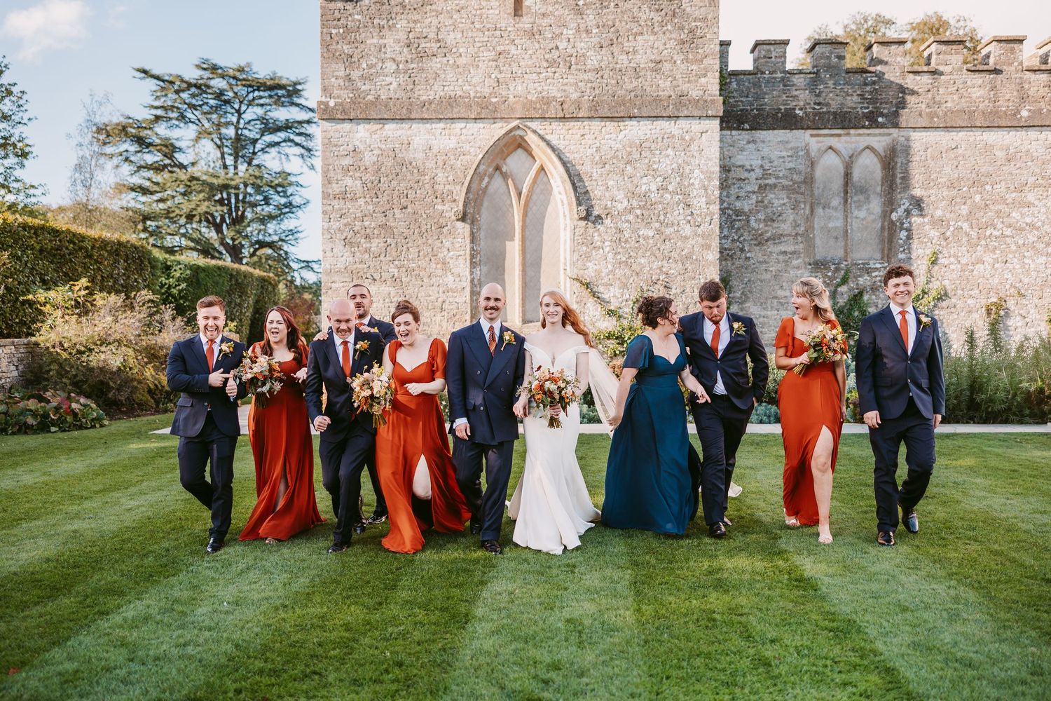 Bride in white gown walks with groom, groomsmen and bridesmaids in rust and teal dresses across a green lawn before a historic stone chapel.