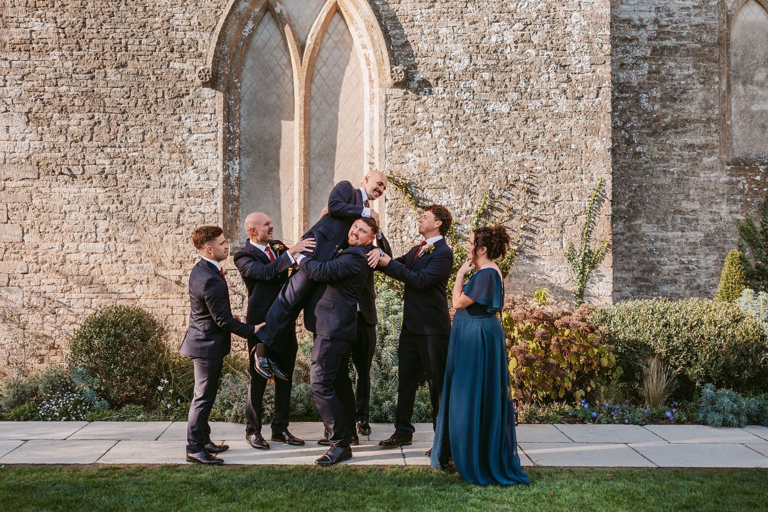 Groomsmen in dark suits playfully lift a groom in front of a stone church while a bridesmaid in a teal gown watches.