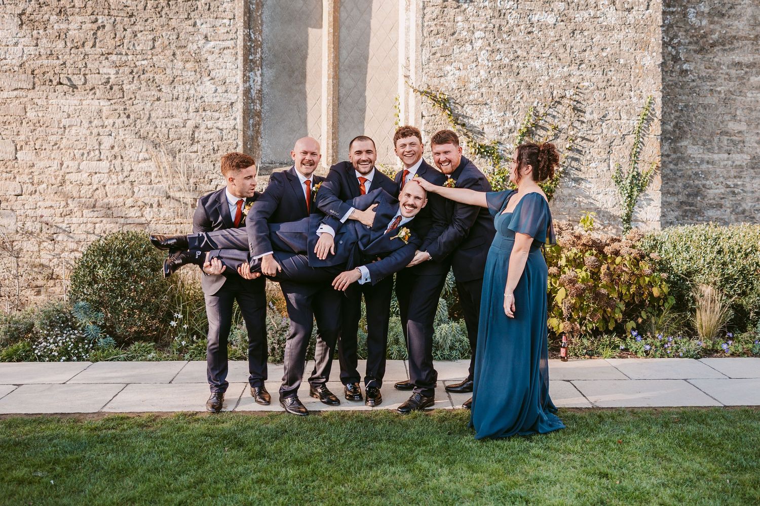 Groomsmen in dark suits laugh while lifting a groom horizontally in a playful pose beside a woman in a teal dress on a stone patio.