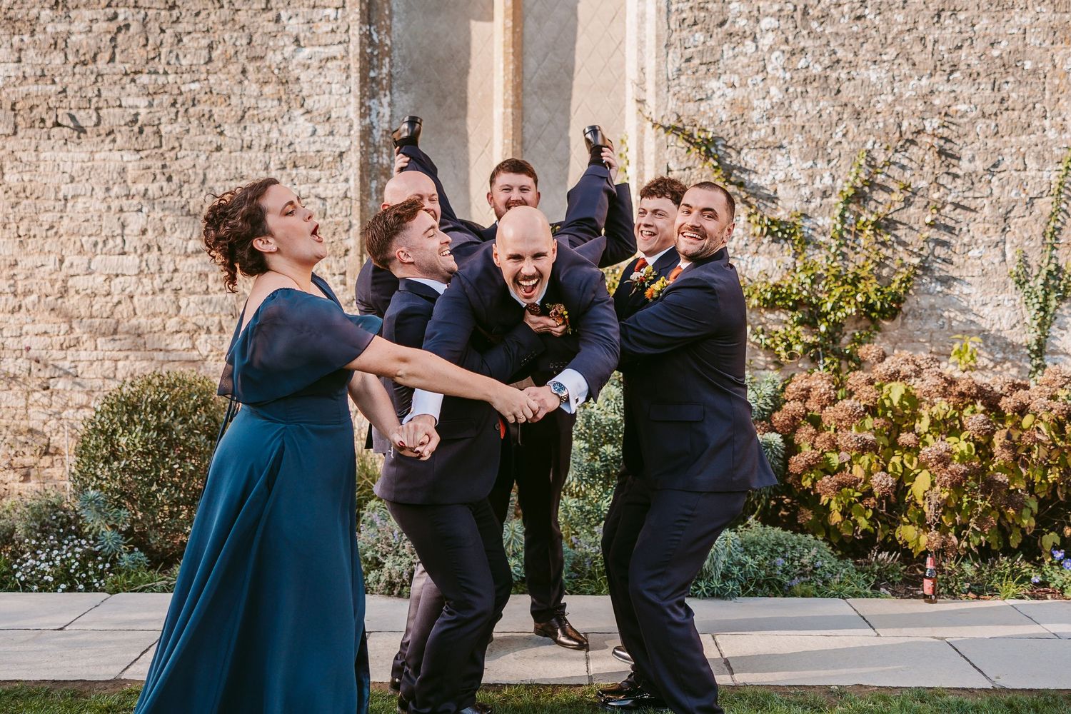 Playful groomsmen in dark suits lift a laughing bridesmaid in a teal gown outside a stone building with flowering shrubs at a wedding.