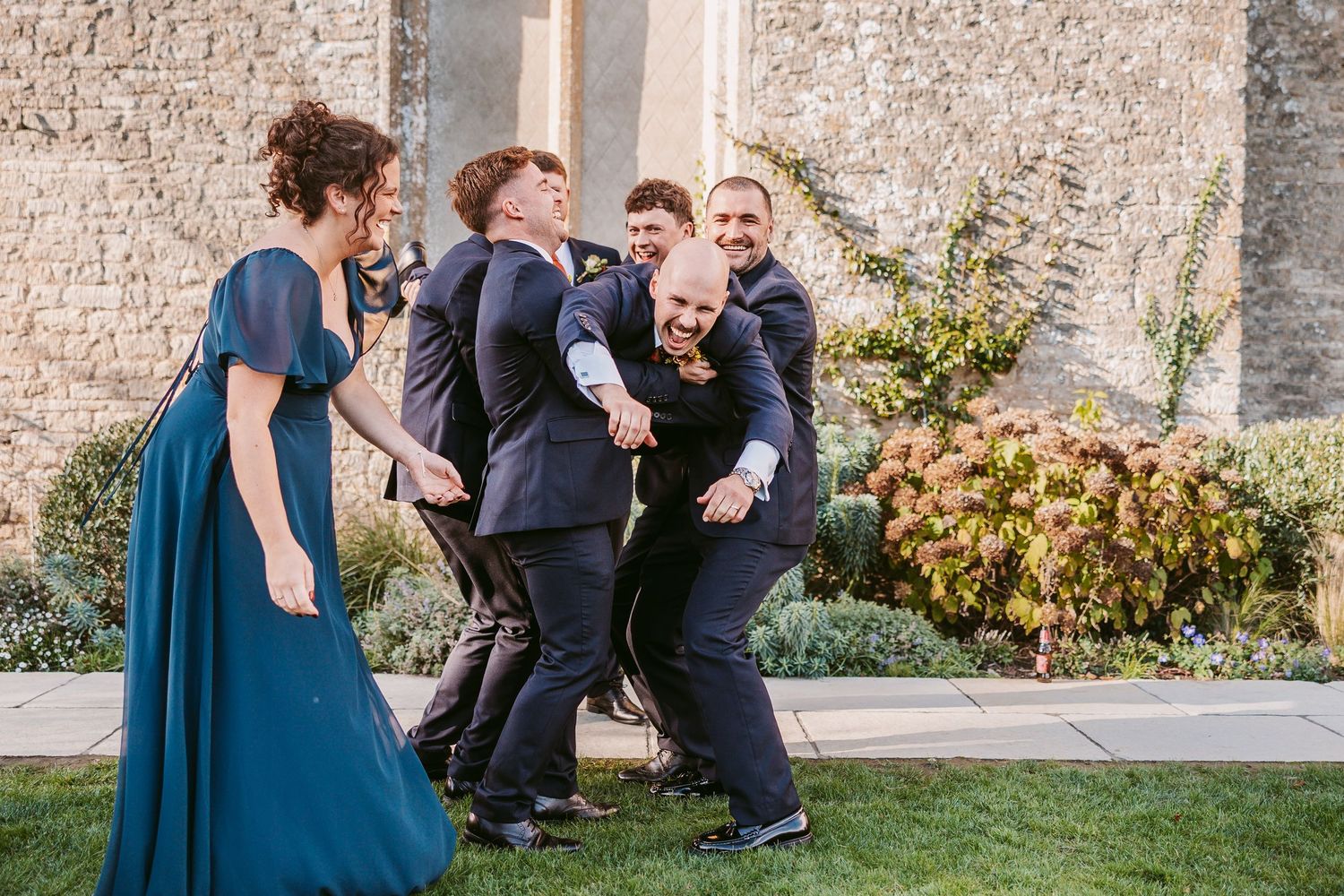 Groomsmen in dark suits laugh and huddle together on a lawn beside a woman in a teal gown at an outdoor stone-wall wedding venue.