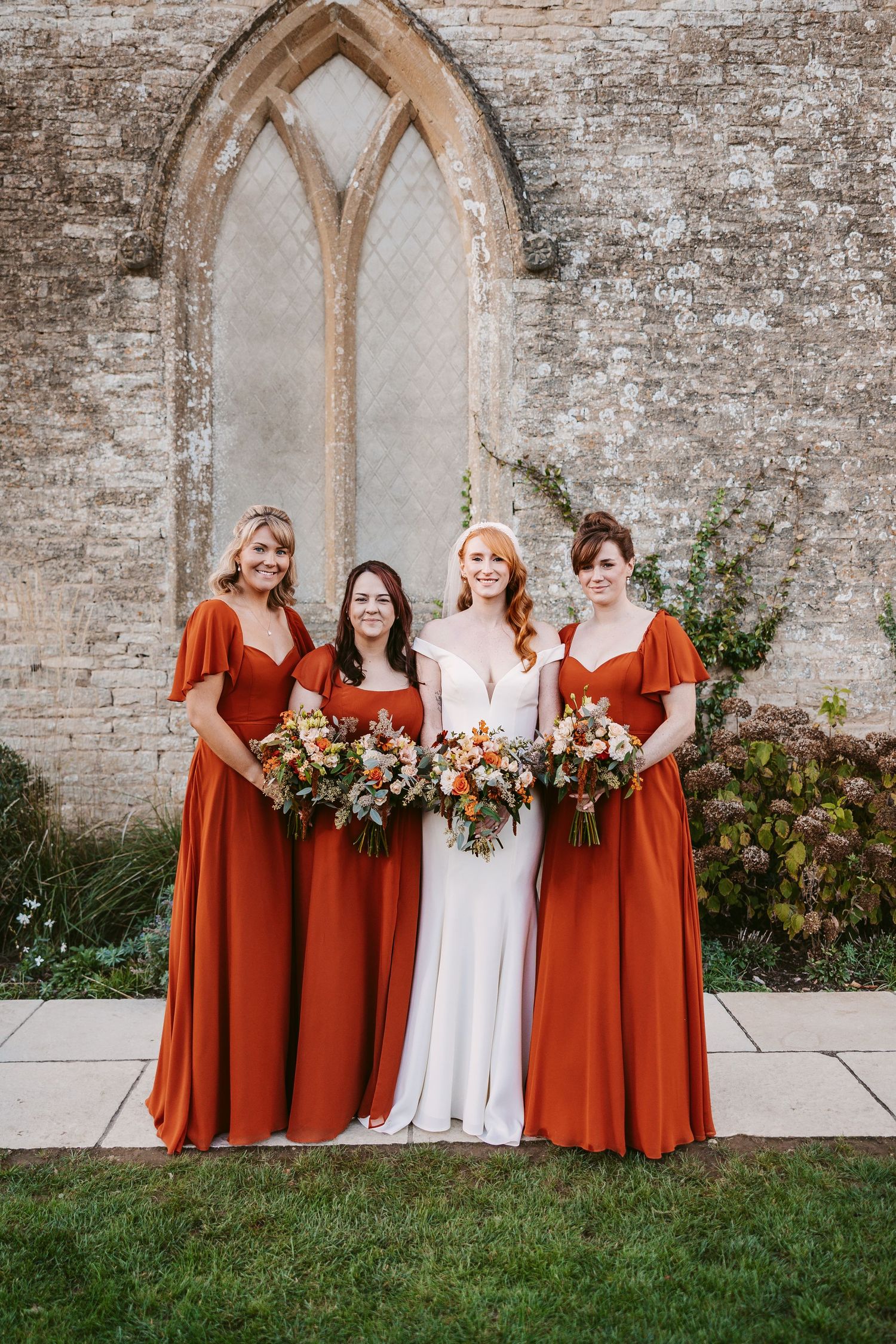 Bride in a white gown stands with three bridesmaids in rust dresses holding autumn bouquets before a stone church window.