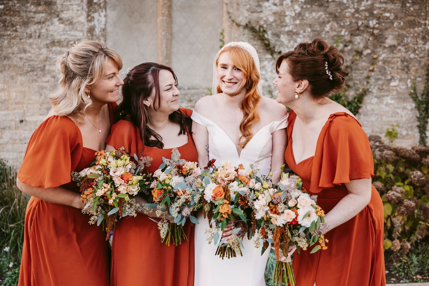 Bride in a white dress laughs with three bridesmaids in rust orange dresses, all holding autumn bouquets beside a stone wall.