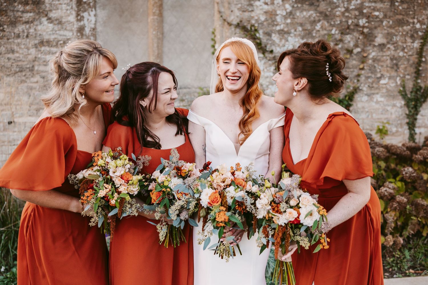 Smiling bride in a white gown stands among three bridesmaids in rust orange dresses, each holding colorful autumn wedding bouquets by a ston