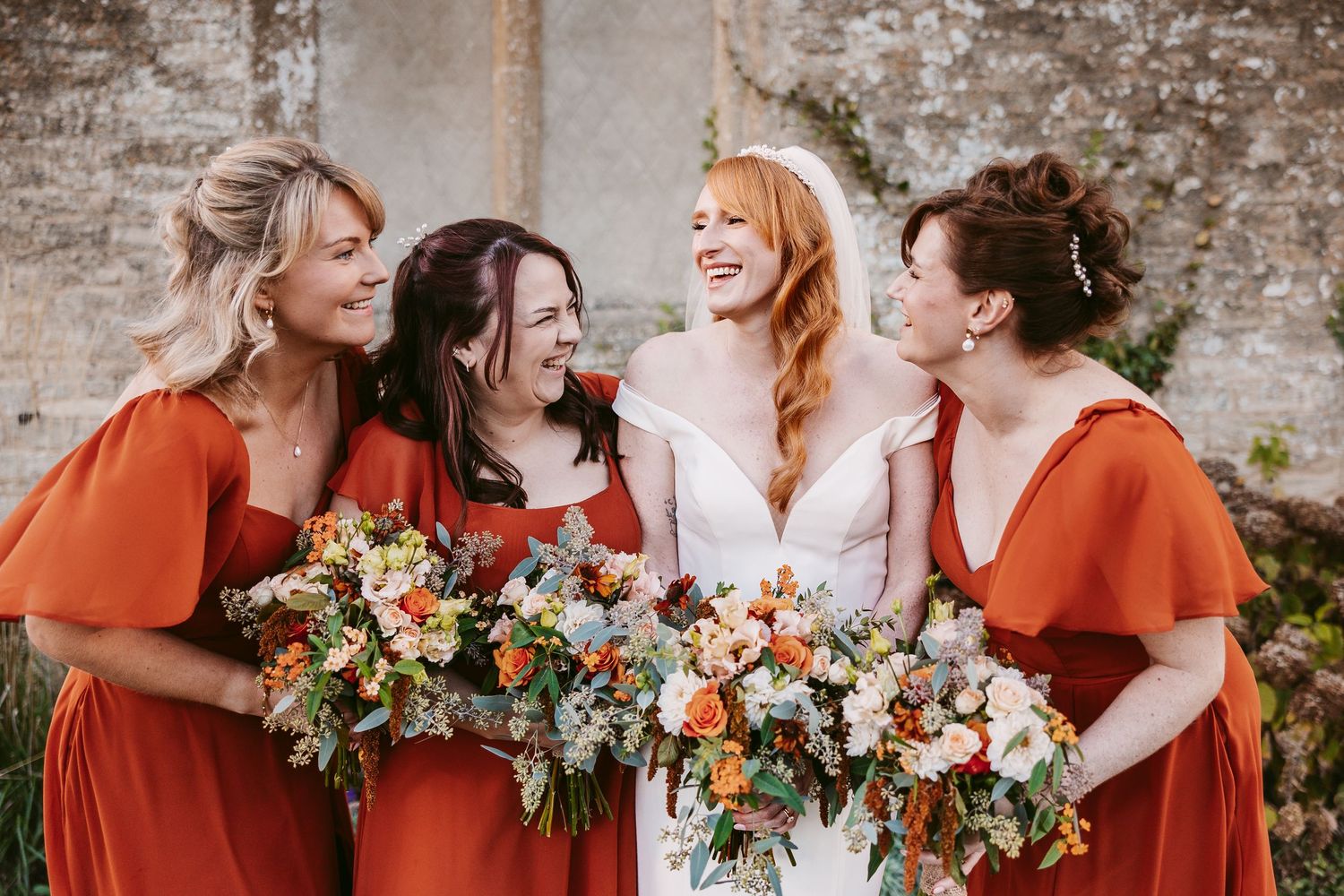 Bride laughs with three bridesmaids in rust orange dresses, all holding colorful autumn wedding bouquets in front of an old stone wall.