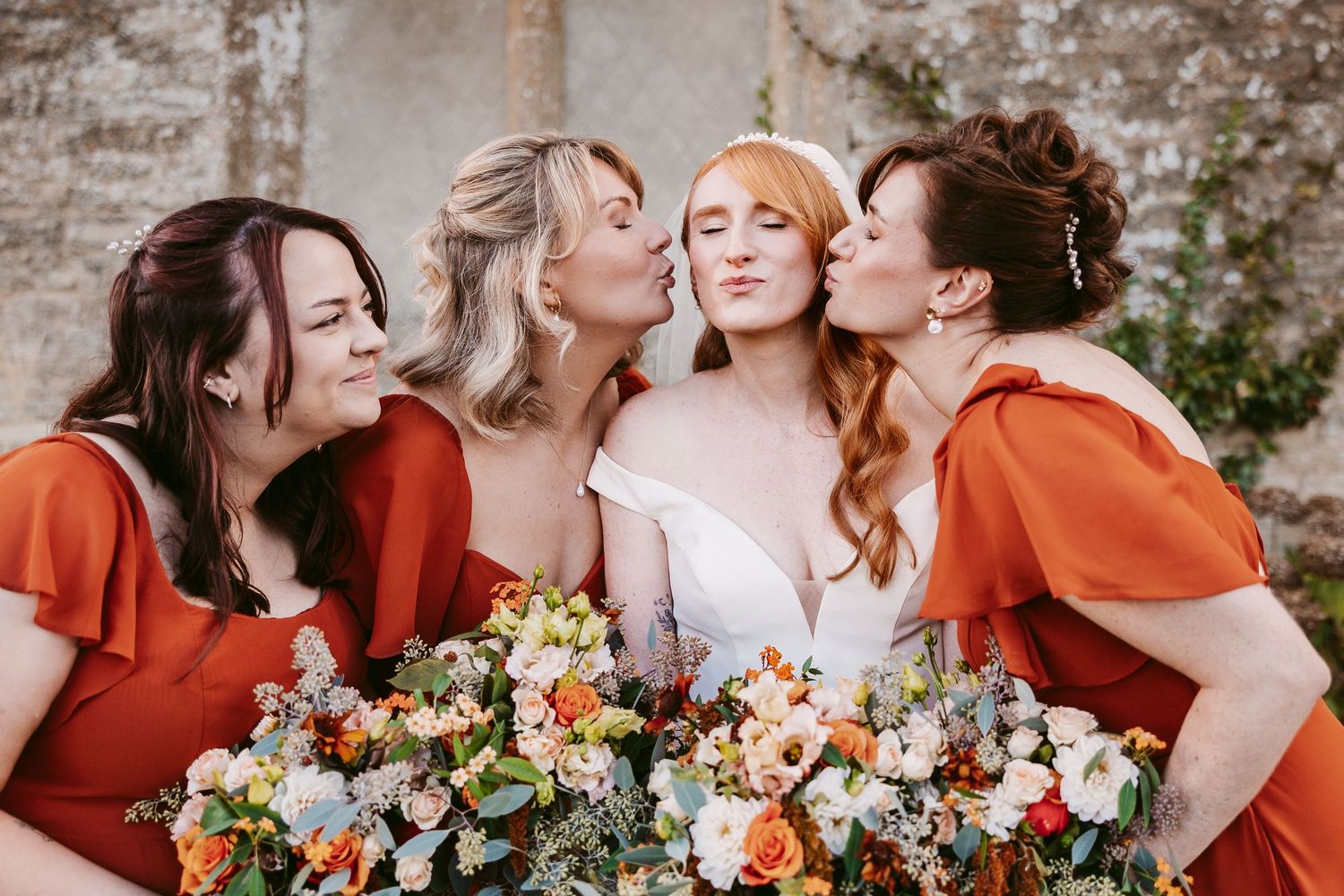 Bride in white and three bridesmaids in rust orange dresses lean in for a kiss on her cheeks beside a colorful wedding bouquet.