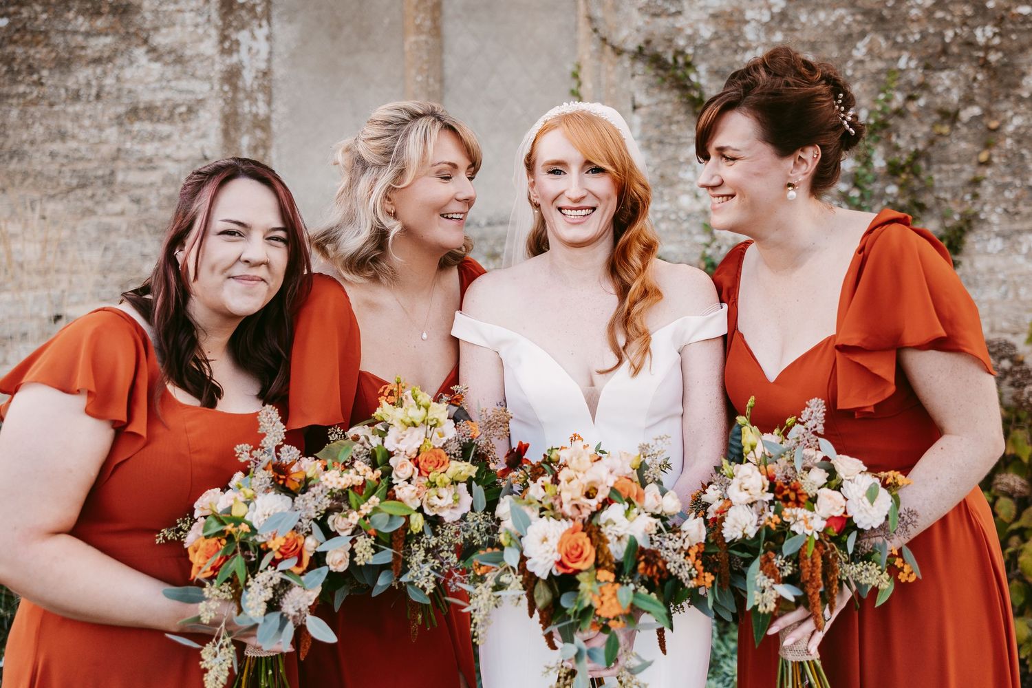 Bride in an off-shoulder white dress laughs with three bridesmaids in rust orange dresses, each holding colorful floral bouquets.