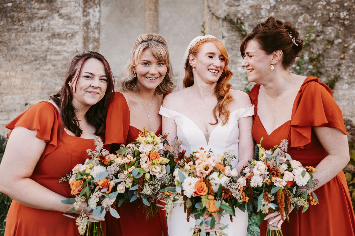 A joyful bride in a strapless white gown poses with four bridesmaids in burnt orange dresses, each holding a lush floral bouquet outdoors.