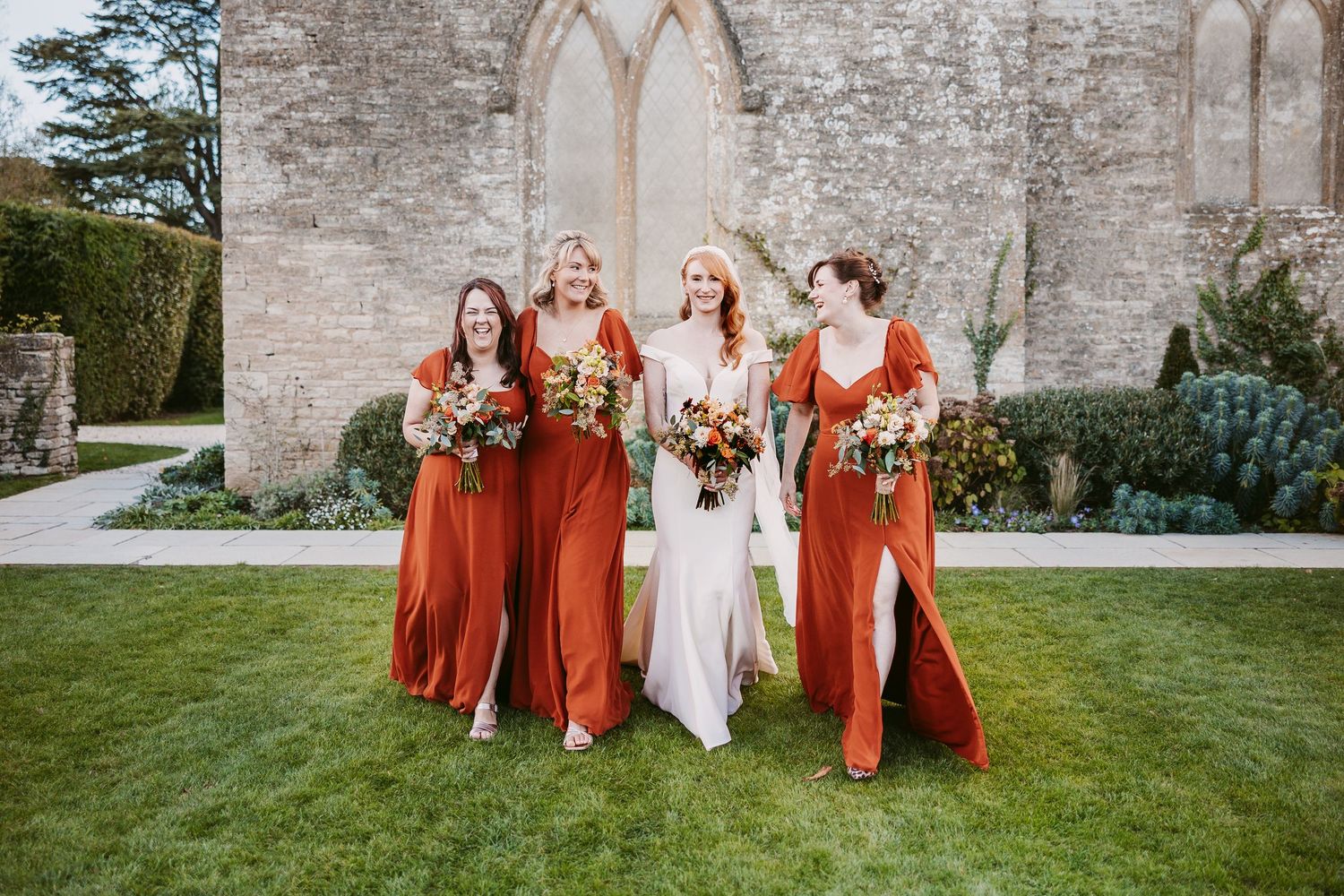Bride in a white gown walks with three bridesmaids in rust orange dresses, holding bouquets on a lawn before a historic stone church.