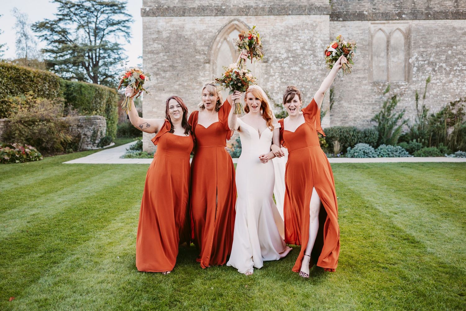 Bride in a white gown and three bridesmaids in rust orange dresses raise bouquets on a green lawn outside a stone wedding venue.