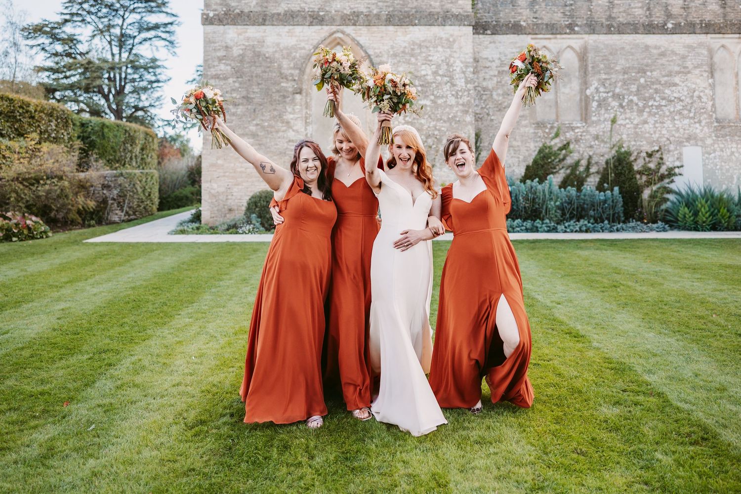 Bride and three bridesmaids in rust orange dresses cheer with raised bouquets on a sunlit lawn beside a stone building.