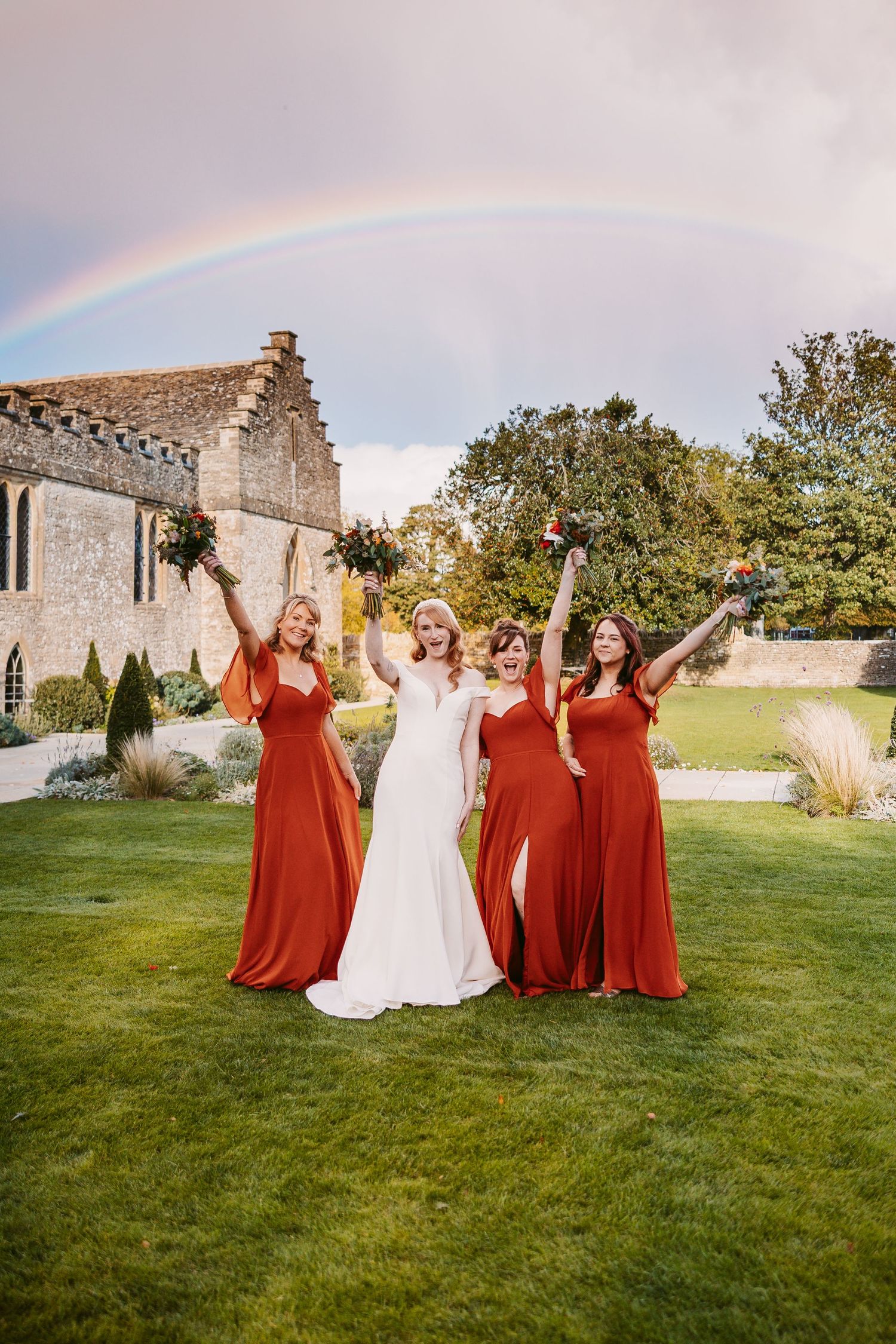 Bride and three bridesmaids in rust dresses cheer with bouquets on a manicured lawn beneath a rainbow at a historic stone wedding venue.