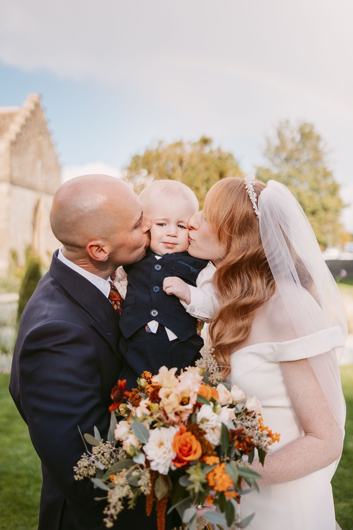 Groom and bride in a veil kiss their baby son, who wears a waistcoat, while holding an autumn bouquet outdoors.