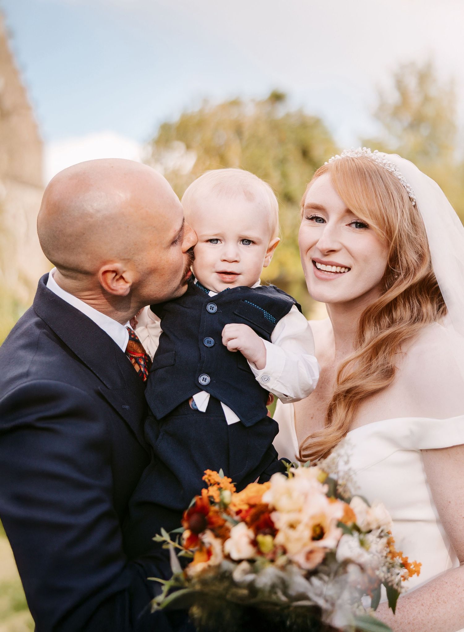 A smiling bride in a veil holds an orange bouquet while a groom kisses their baby boy, dressed in a navy waistcoat, outdoors.