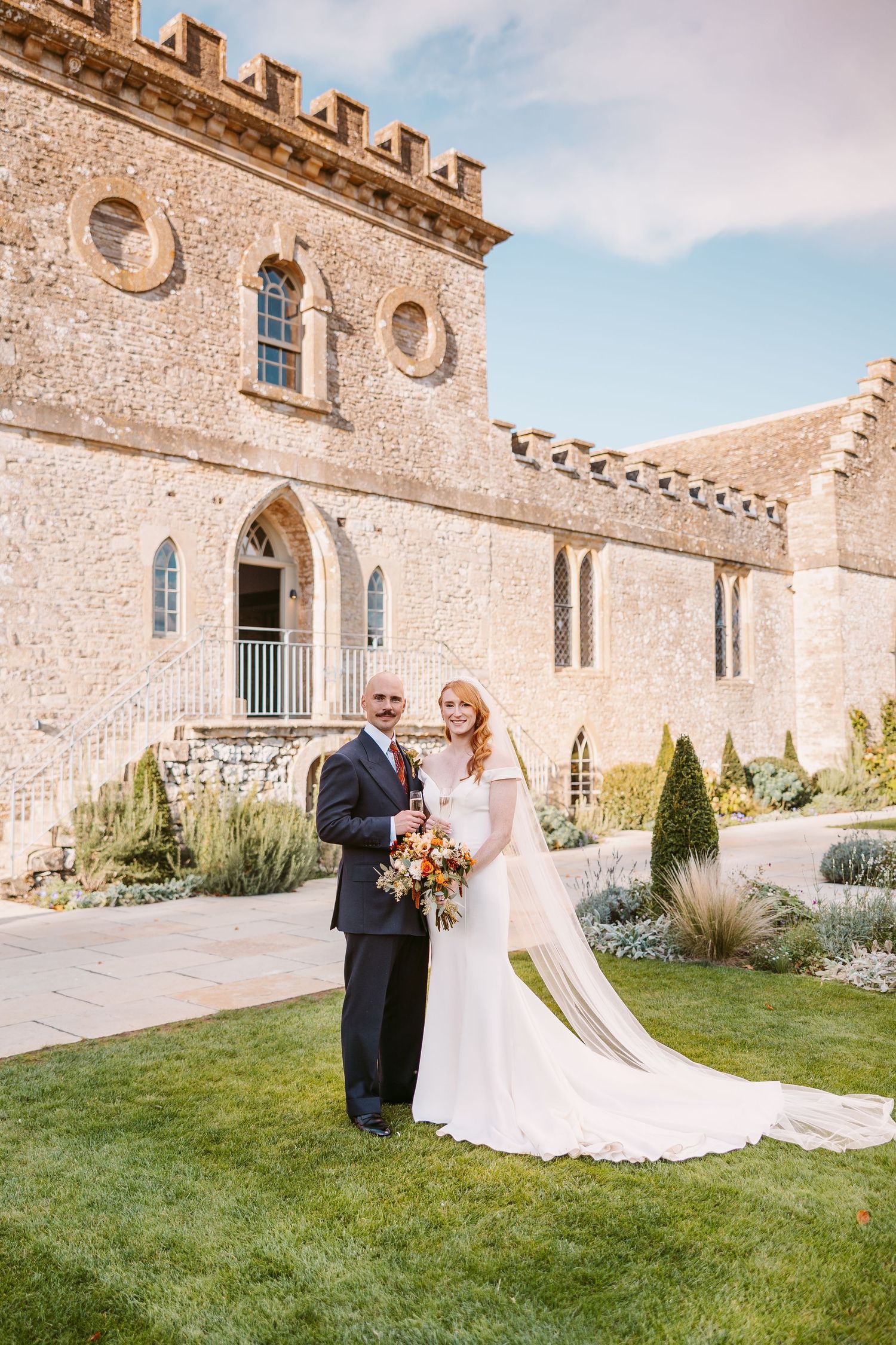 Bride in a fitted white gown with a long veil and groom in a navy suit hold a bouquet on the lawn before a castle-style stone wedding venue.