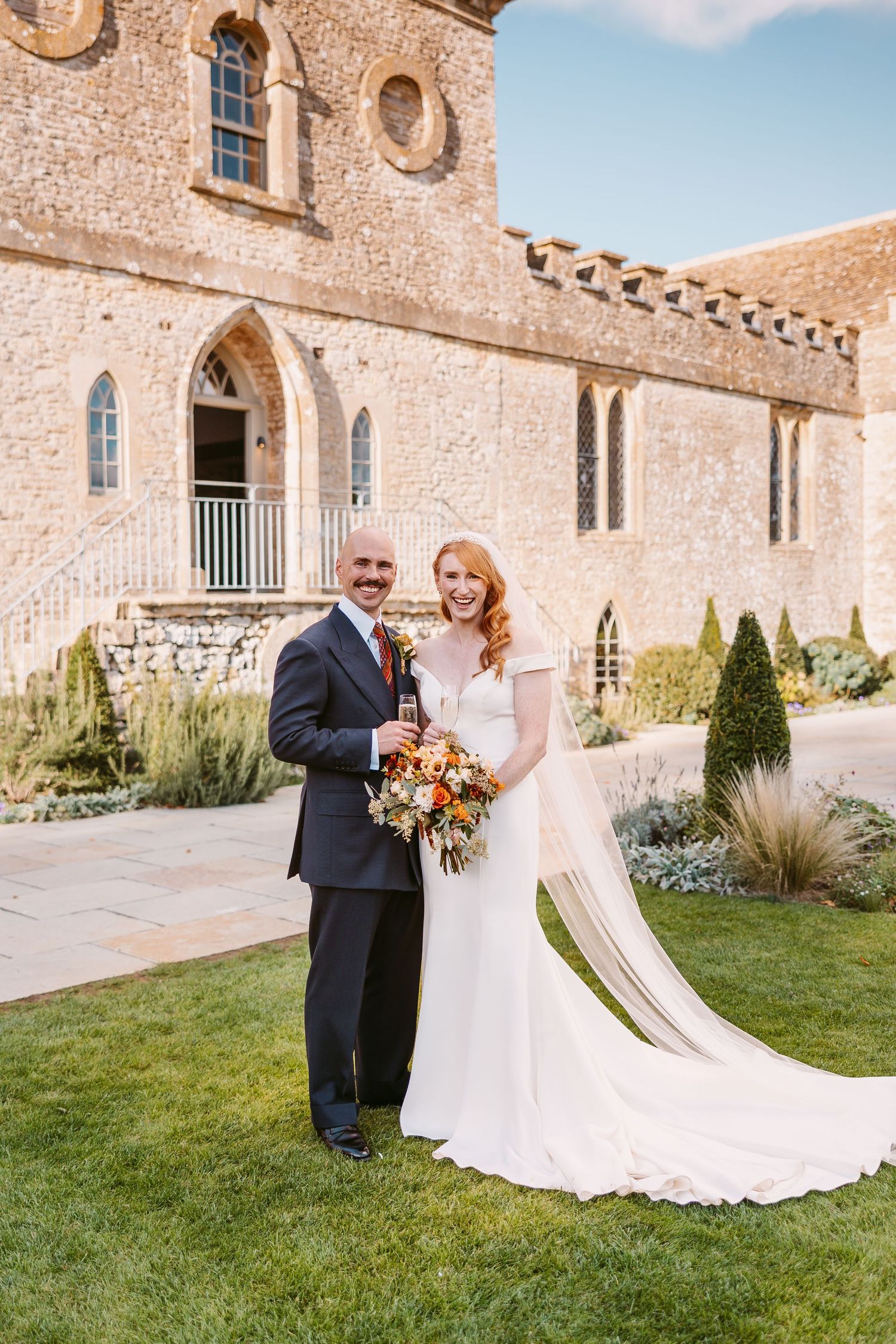 Bride in a white gown and groom in a dark suit hold champagne and a bouquet in front of a historic stone wedding venue.