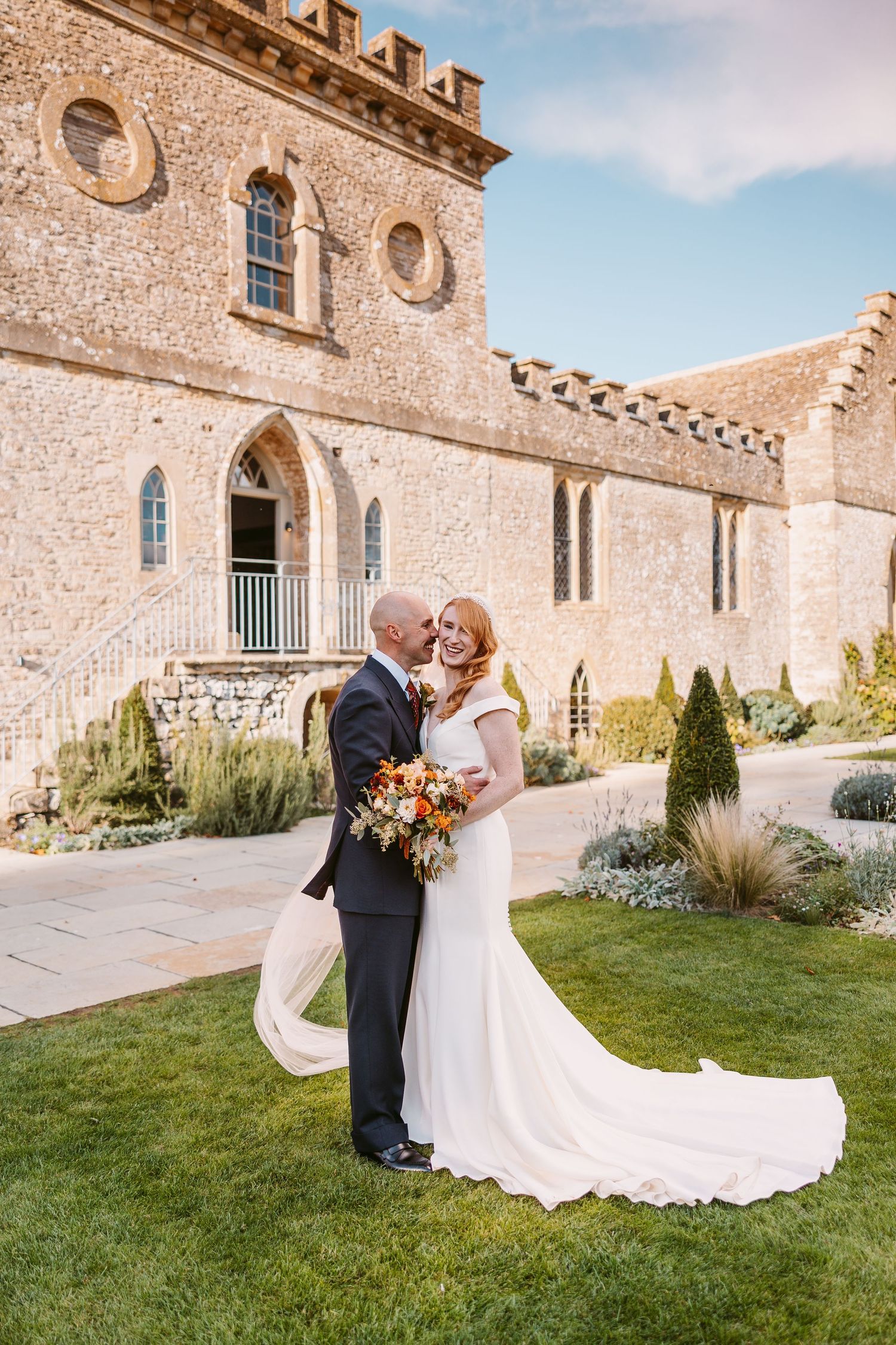 Bride in a white gown and groom in a suit share a kiss on the lawn in front of a historic stone castle wedding venue.