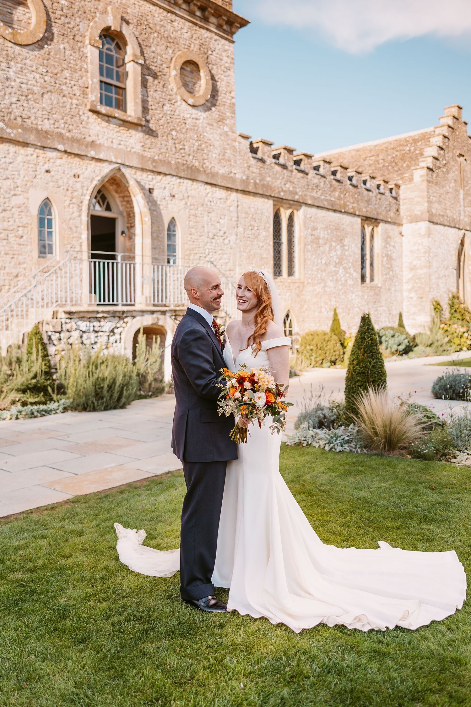 Smiling bride holding an orange bouquet embraces her groom on a green lawn beside a medieval stone building at their wedding.