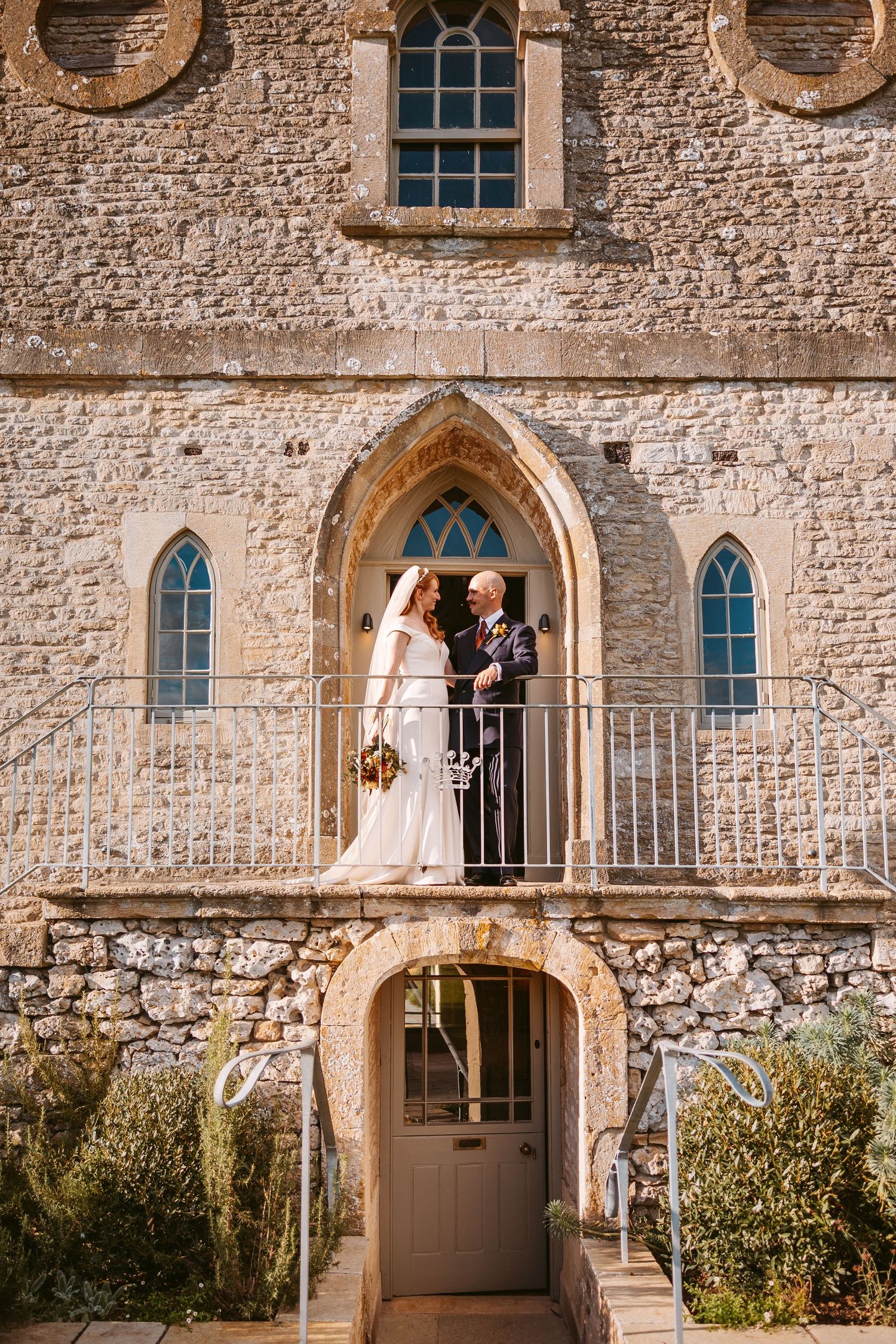 Newlyweds stand together on the balcony of an arched stone building with the bride's long veil flowing down the steps.