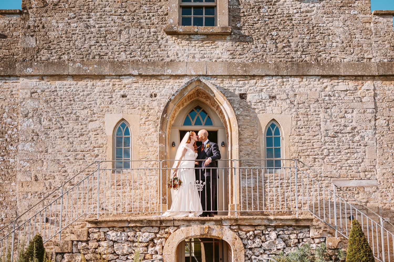 A bride and groom embrace beneath a pointed Gothic arch at the top of stone steps outside a historic stone wedding venue.