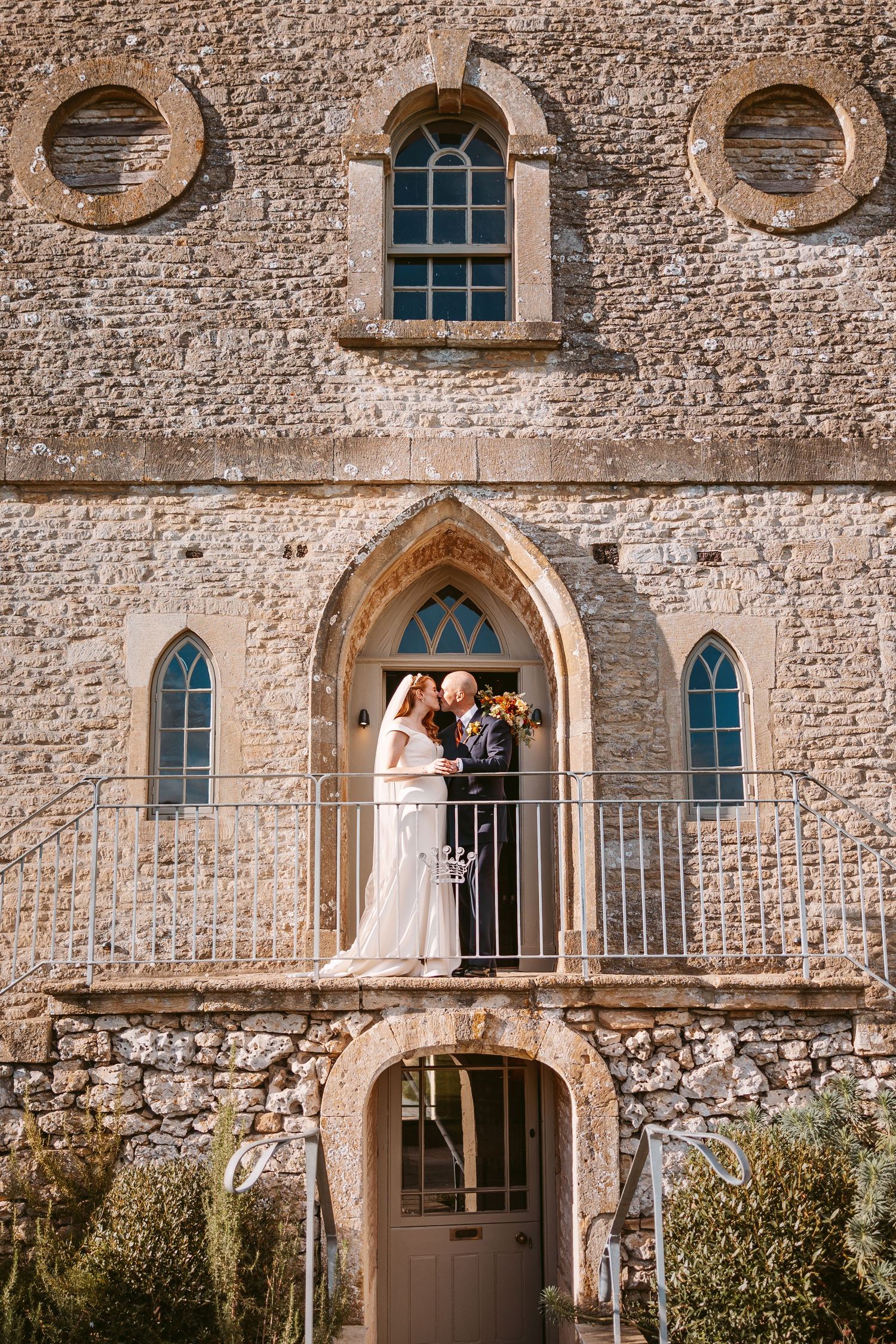 Bride and groom share a close moment on the balcony of a historic stone building with arched windows and a metal railing.