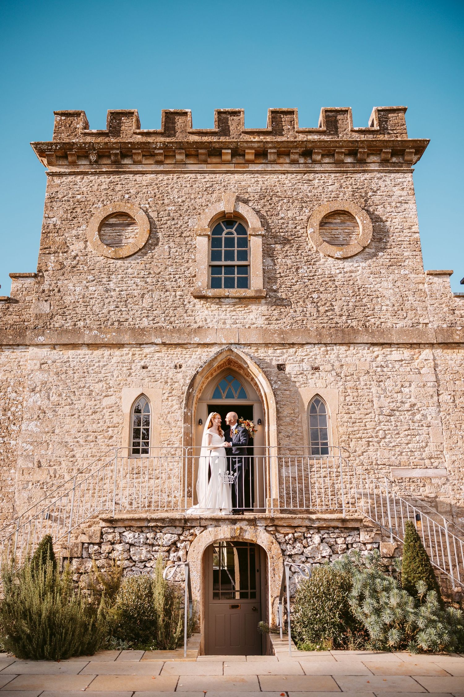 Wedding couple stands on the balcony of a castle-style stone venue with battlements against a clear blue sky.