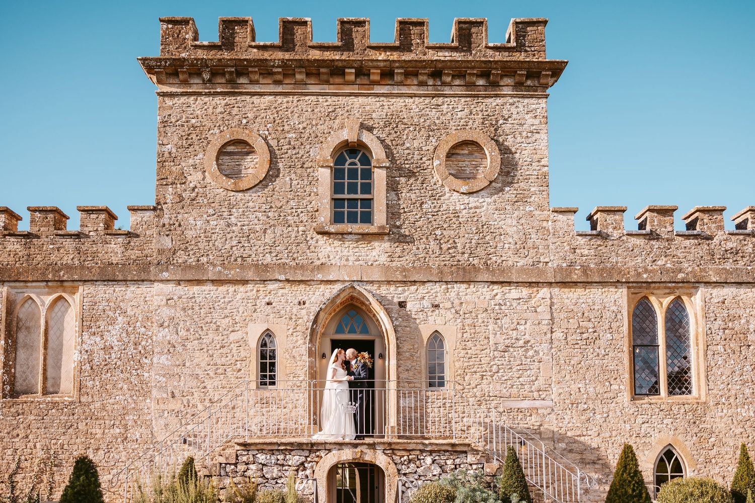 Bride and groom stand in the arched doorway of a crenellated stone castle wedding venue with topiary in the foreground.