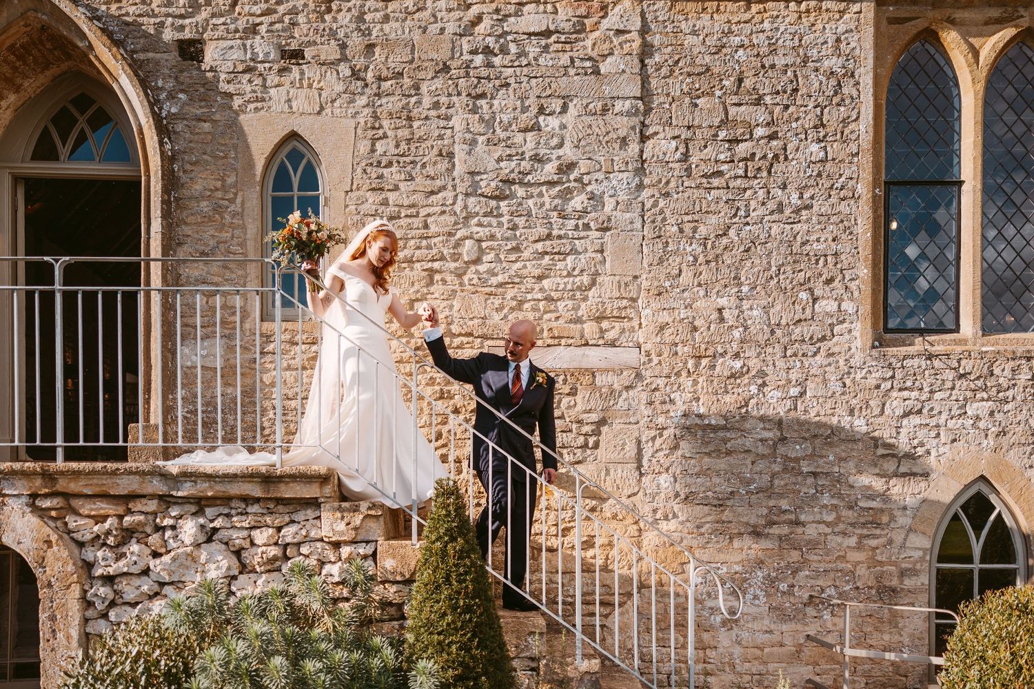 A bride in a flowing white gown holds a bouquet and takes the hand of a groom in a dark suit as they descend stone steps.