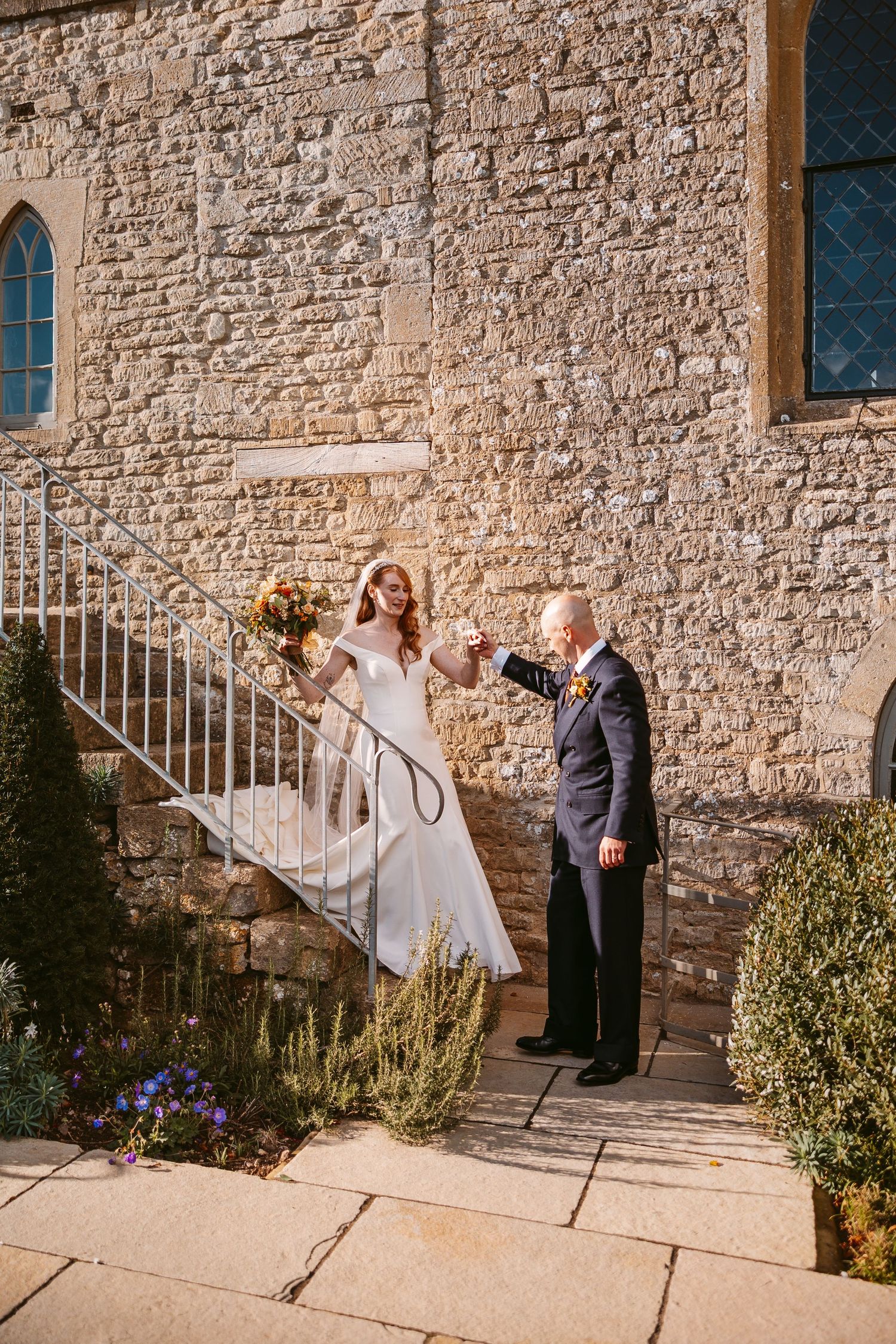 Groom in a navy suit holds the bride's hand as she descends stone steps in a white gown with an orange bouquet.