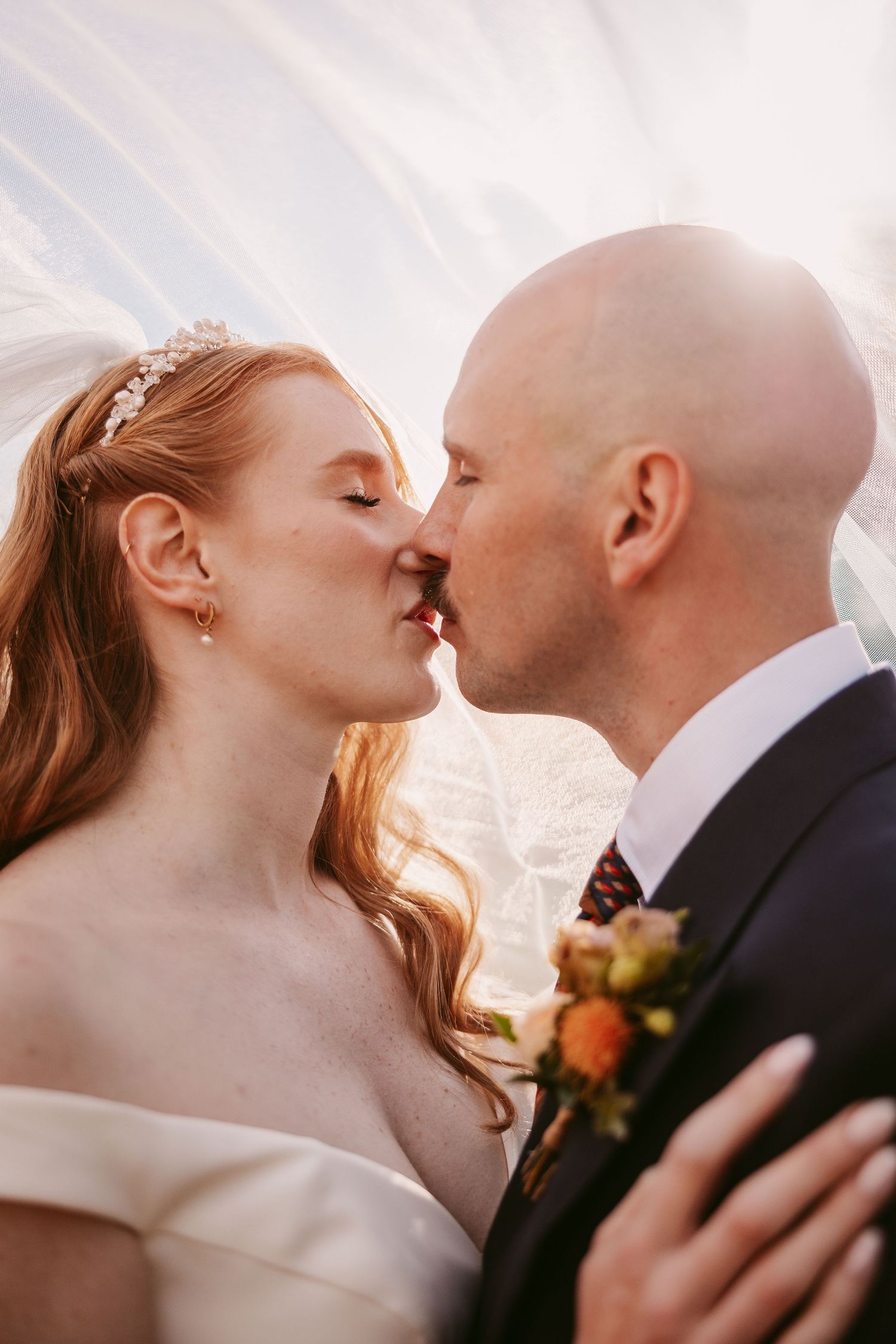 Bride with red hair and a tiara kisses her bald groom in a suit and boutonniere beneath a sheer wedding veil.