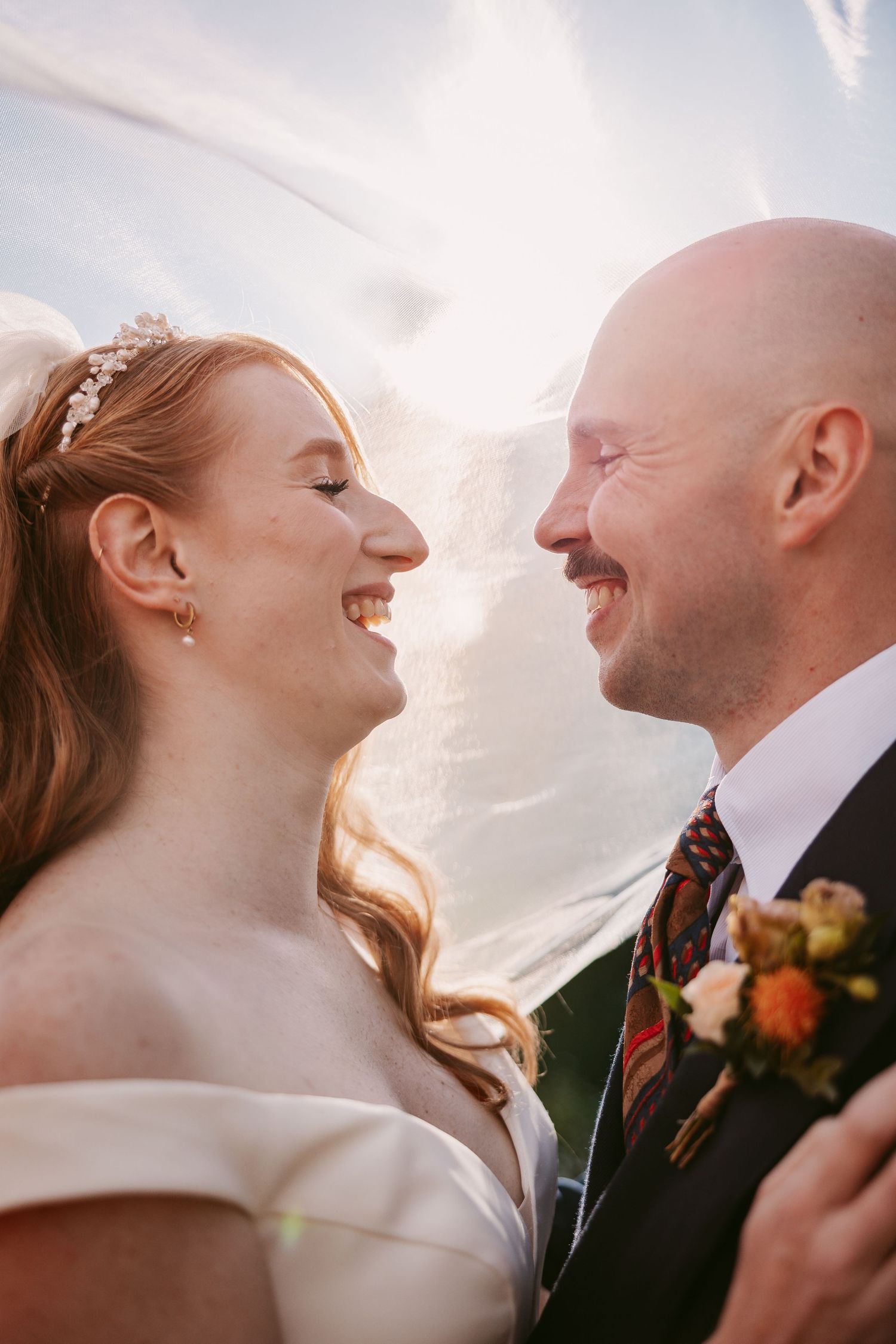 A laughing bride with a floral tiara and a smiling groom in a plaid tie face each other beneath a sheer, sunlit wedding veil.