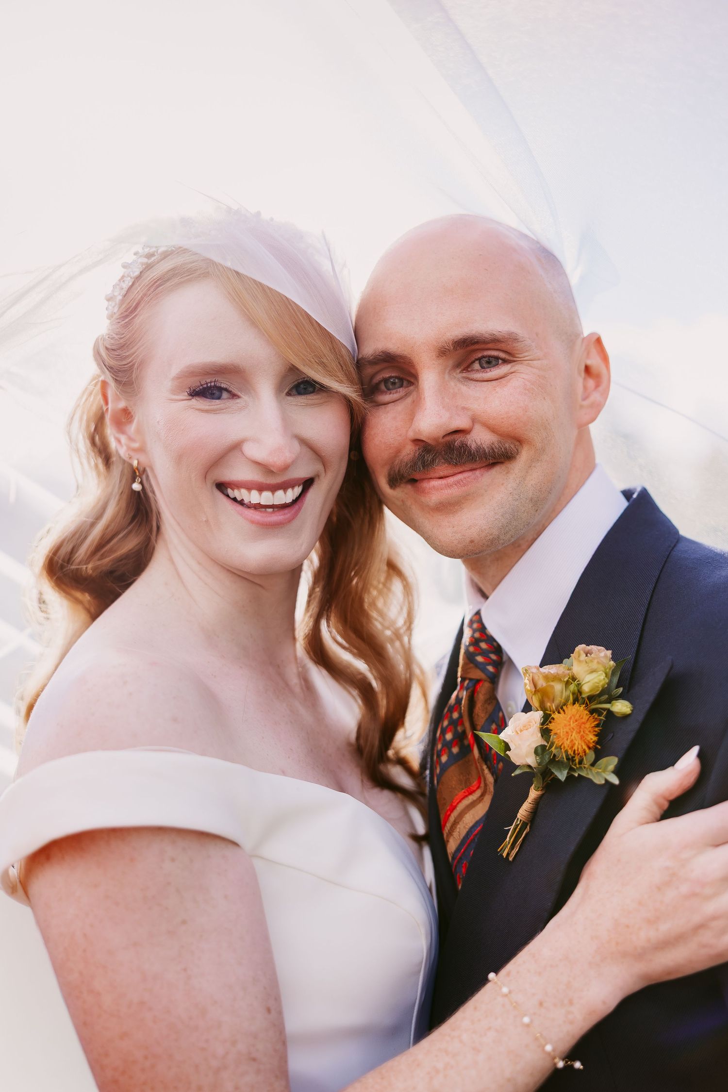Smiling bride in a white off-the-shoulder dress and veil cheek to cheek with her groom in a navy suit and floral boutonniere.