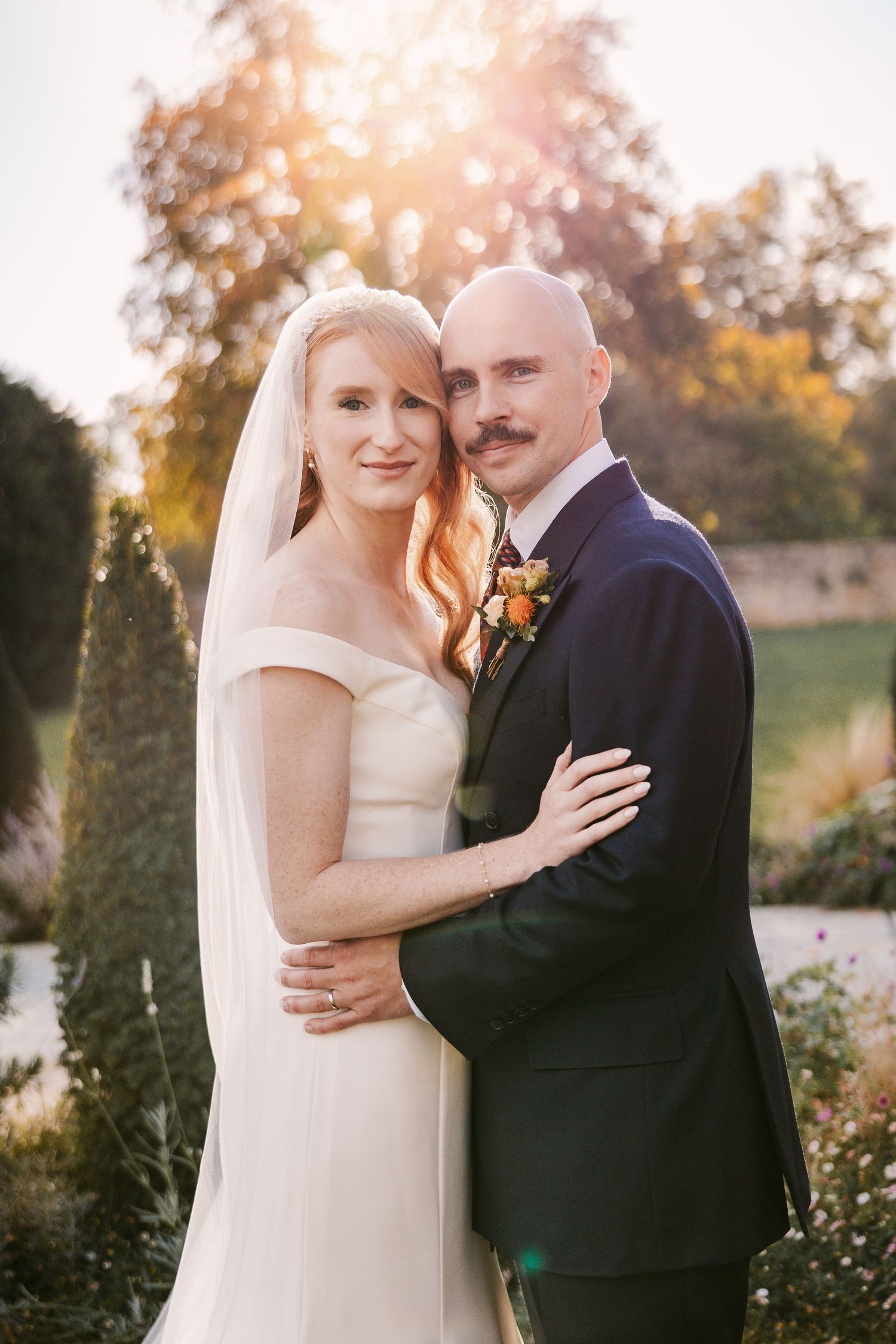 Bride in an off-the-shoulder gown and long veil embraces her groom in a navy suit in a sunlit garden at their wedding.