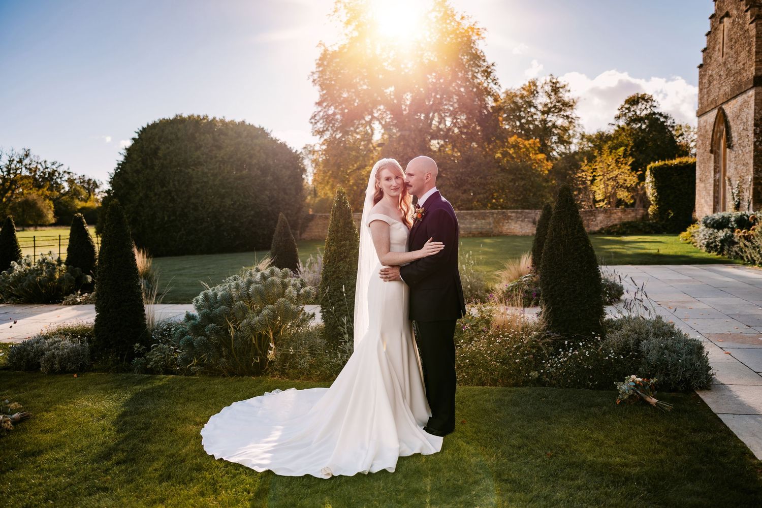 Bride in a white gown and groom embrace on a manicured garden lawn at sunset, framed by topiary and a stone building.