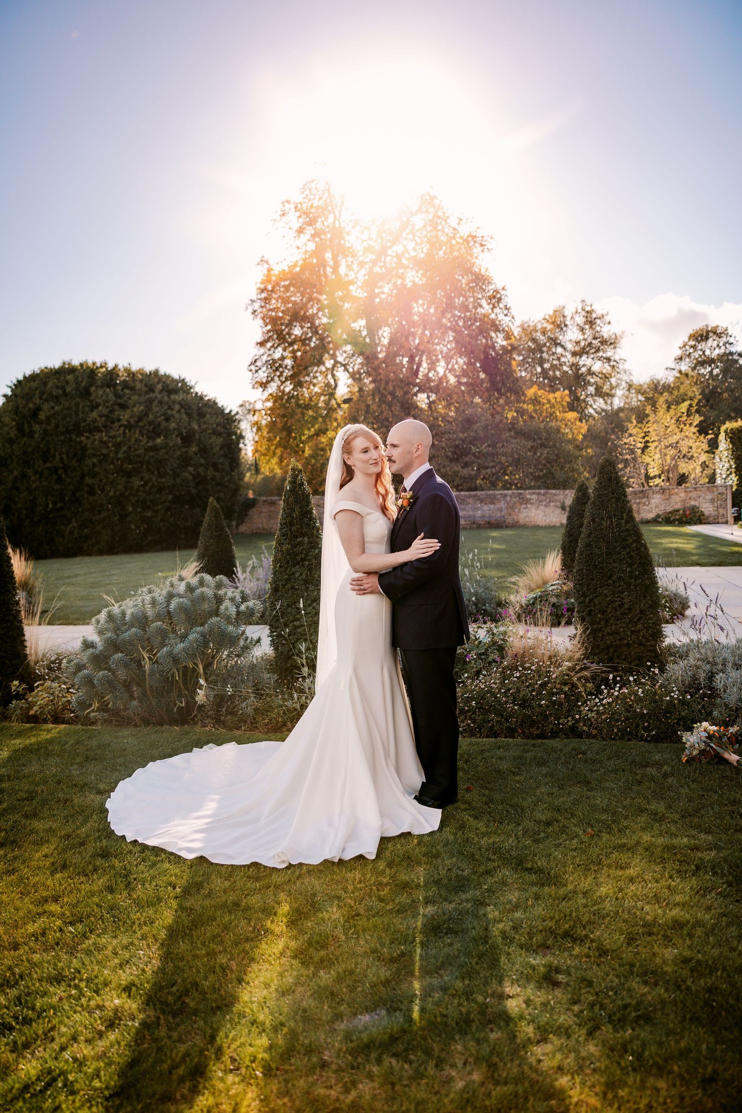 Bride in a long-trained white gown embraces her groom in a sunlit garden with clipped topiary and golden autumn trees.