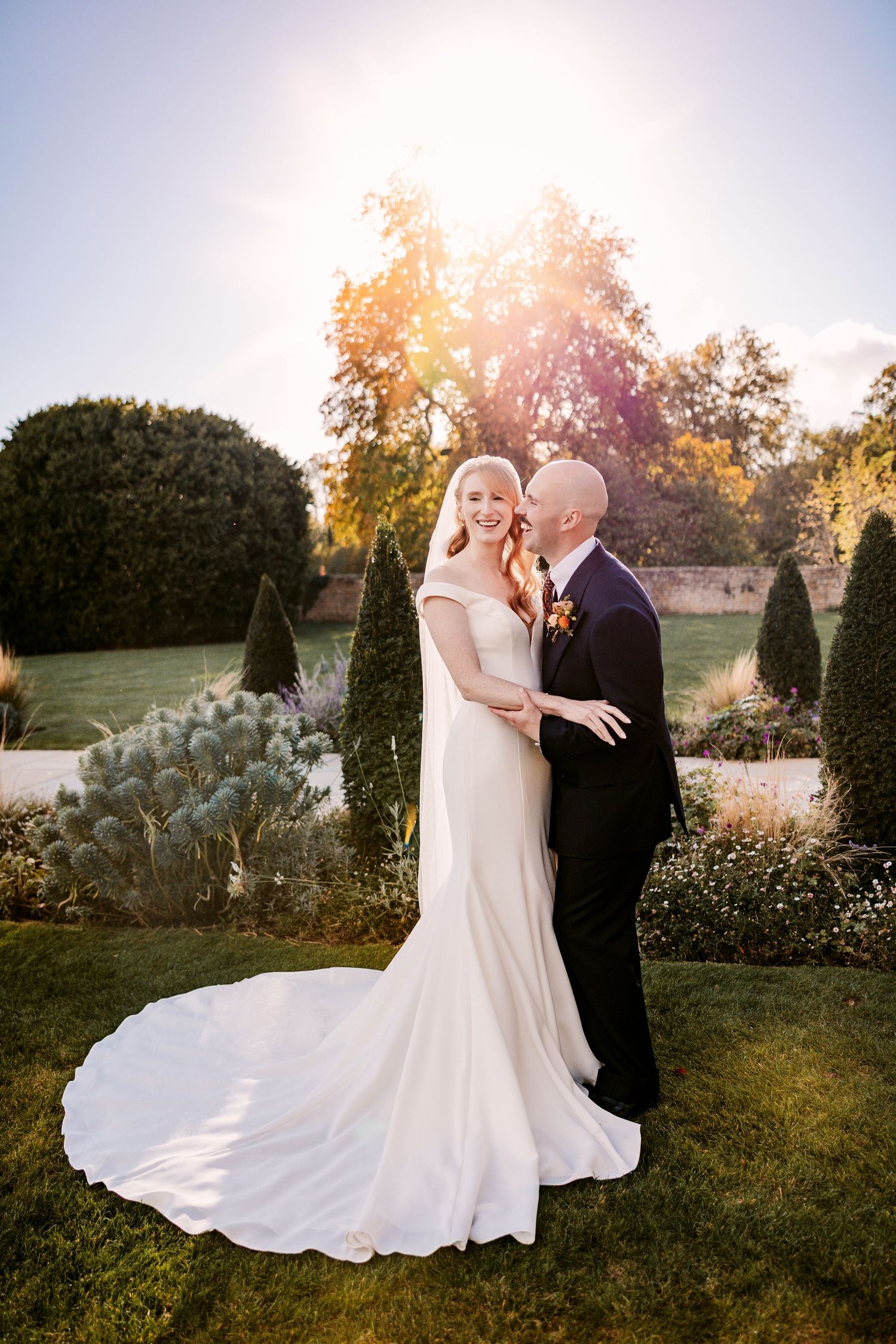 A bride in a long white gown and a groom in a navy suit embrace in a sunlit garden with golden autumn trees behind them.