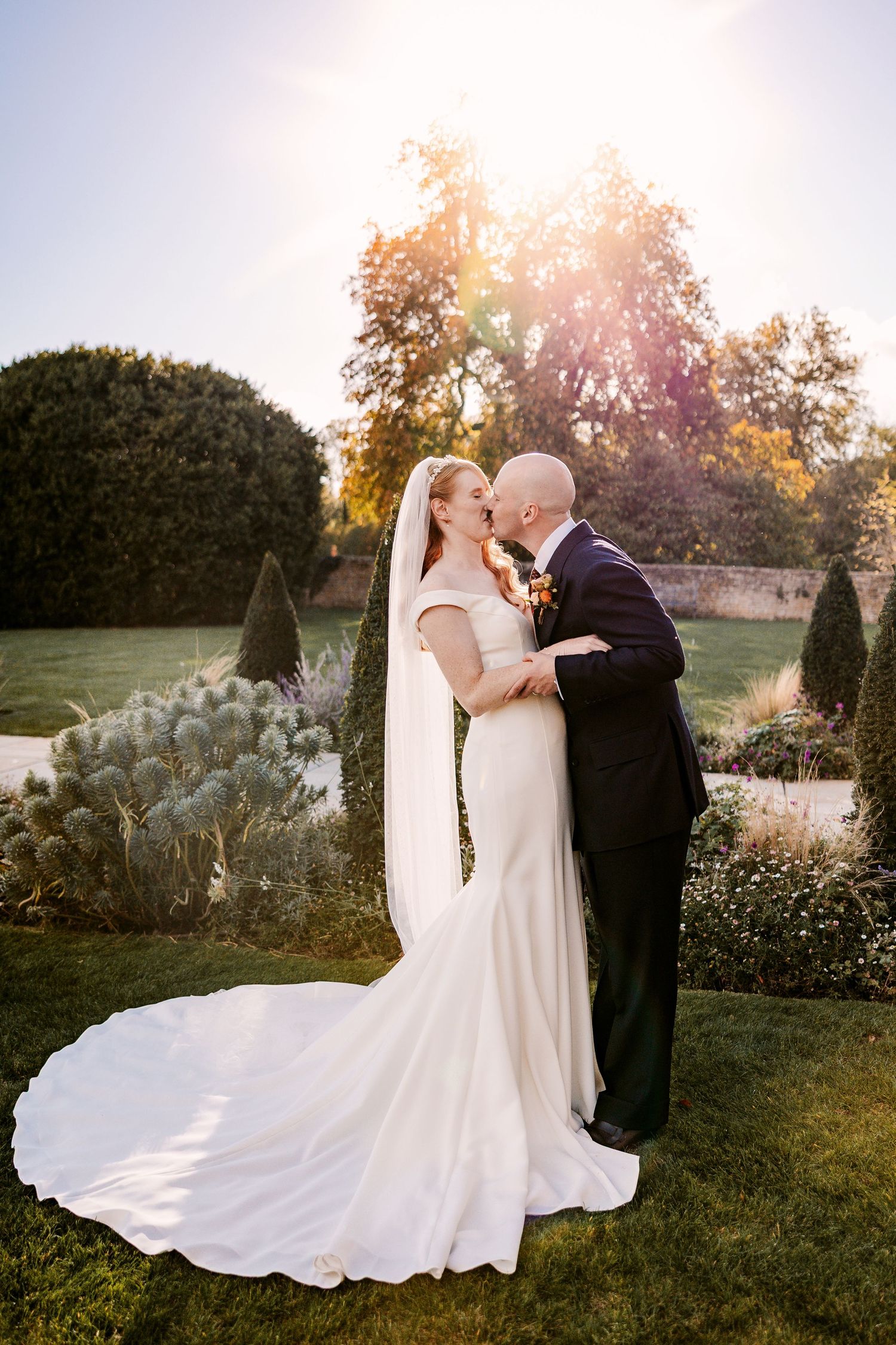 Newlywed couple kisses on a manicured lawn as golden sunlight streams through autumn trees, the bride's long gown train spread behind.