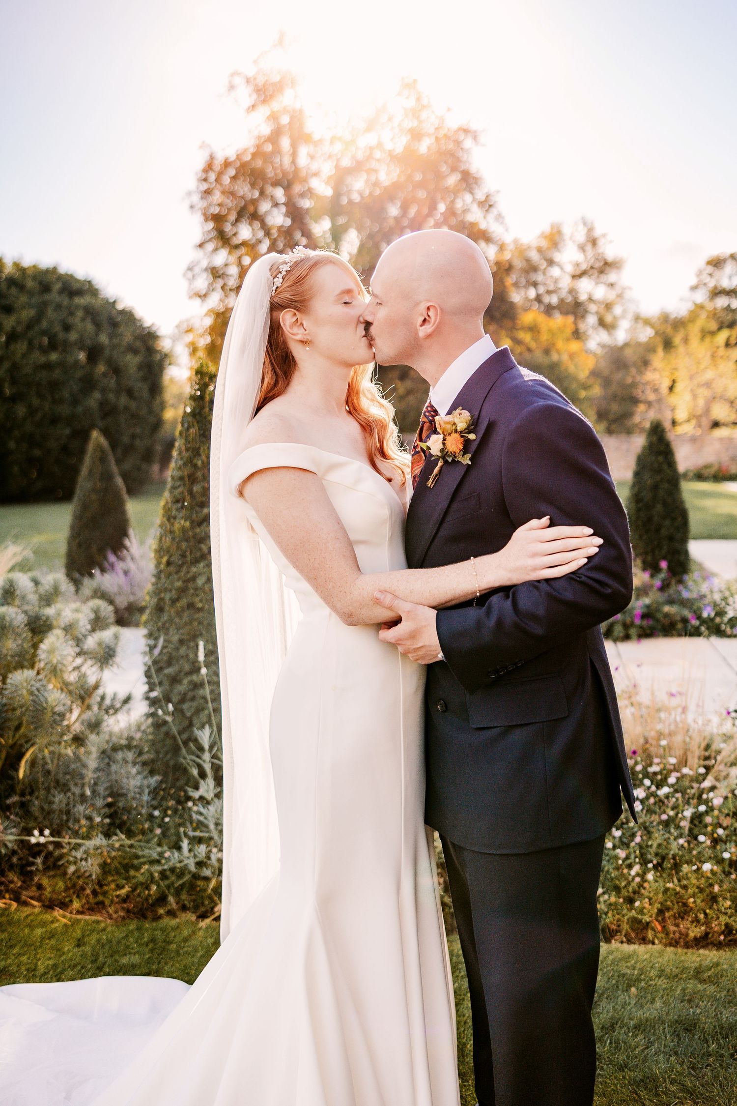 Bride and groom share a kiss in a sunlit garden, surrounded by topiary and autumn trees on their wedding day.