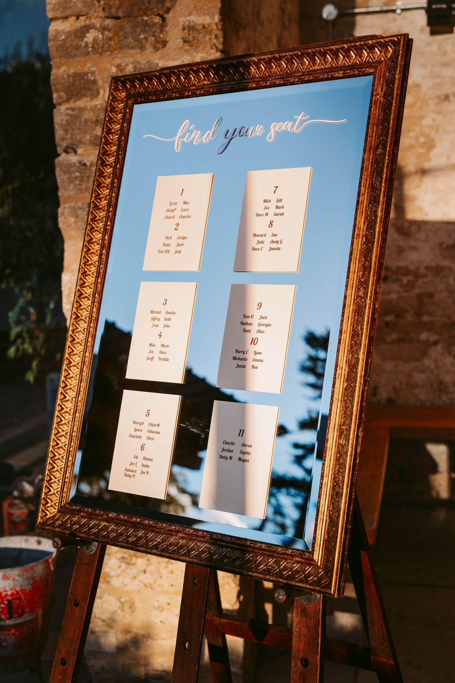 Gold-framed mirror wedding seating chart reading find your seat, with table cards displayed on an easel outdoors.