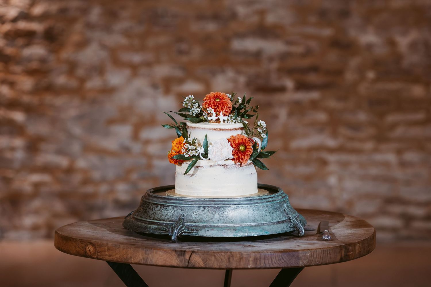 Two-tier white wedding cake decorated with orange flowers and greenery on a rustic table against an exposed brick wall.