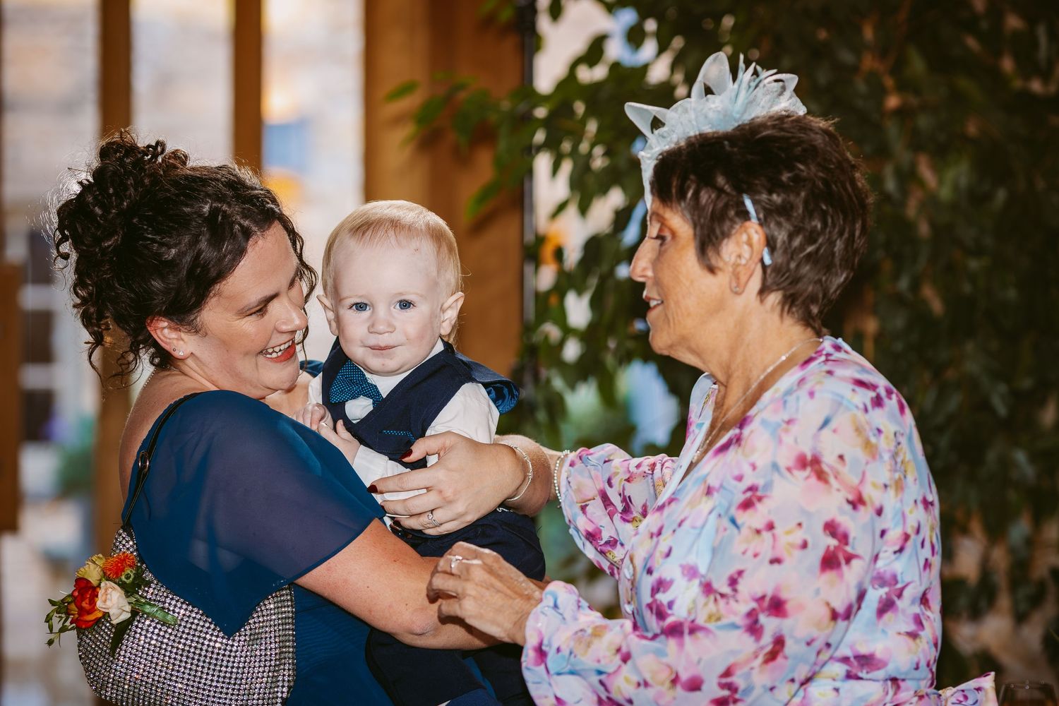 A smiling woman in a navy dress holds a baby boy in a bow tie while an older woman in a floral dress and fascinator gently touches his hand.