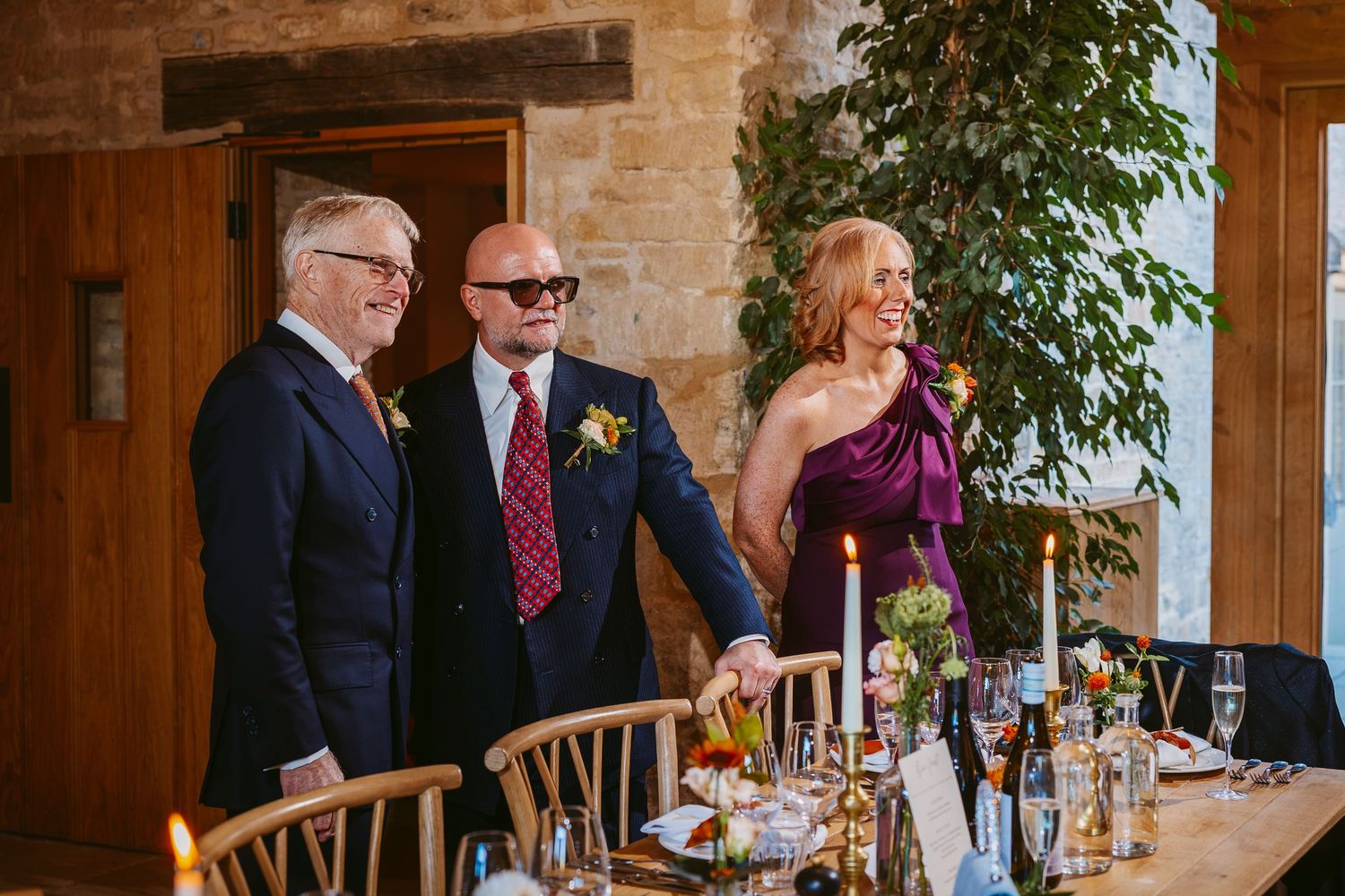 Two men in suits and a woman in a purple dress smile behind a reception table with candles, glasses and flowers.