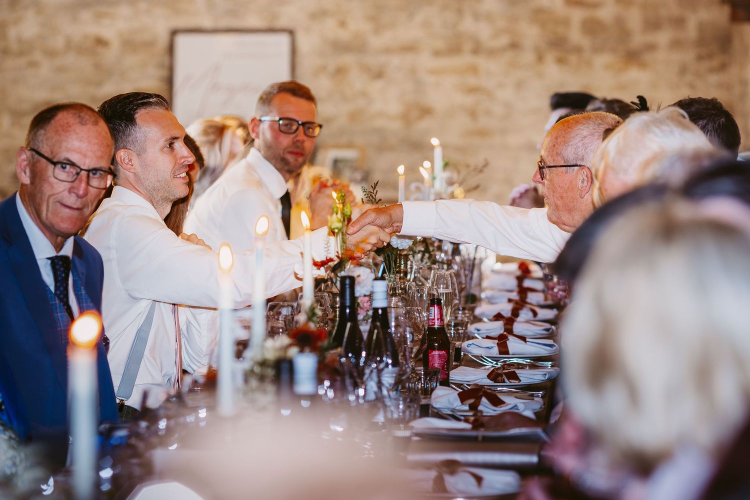 Wedding guests laugh and chat around a candlelit banquet table set with wine bottles, glasses and place settings in a rustic stone venue.