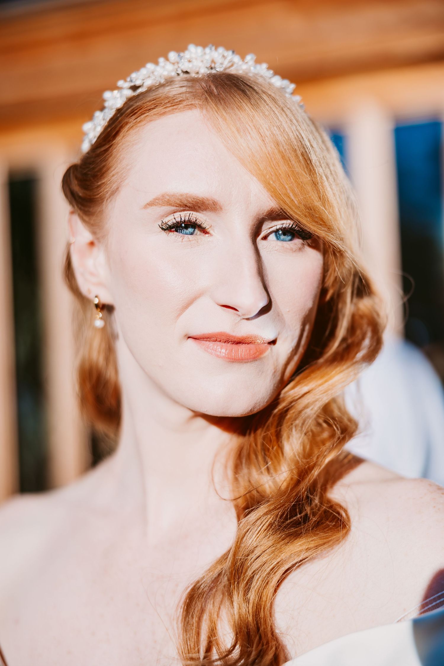 Smiling bride with long red wavy hair wears a pearl tiara and drop earrings, glowing in soft golden sunlight.