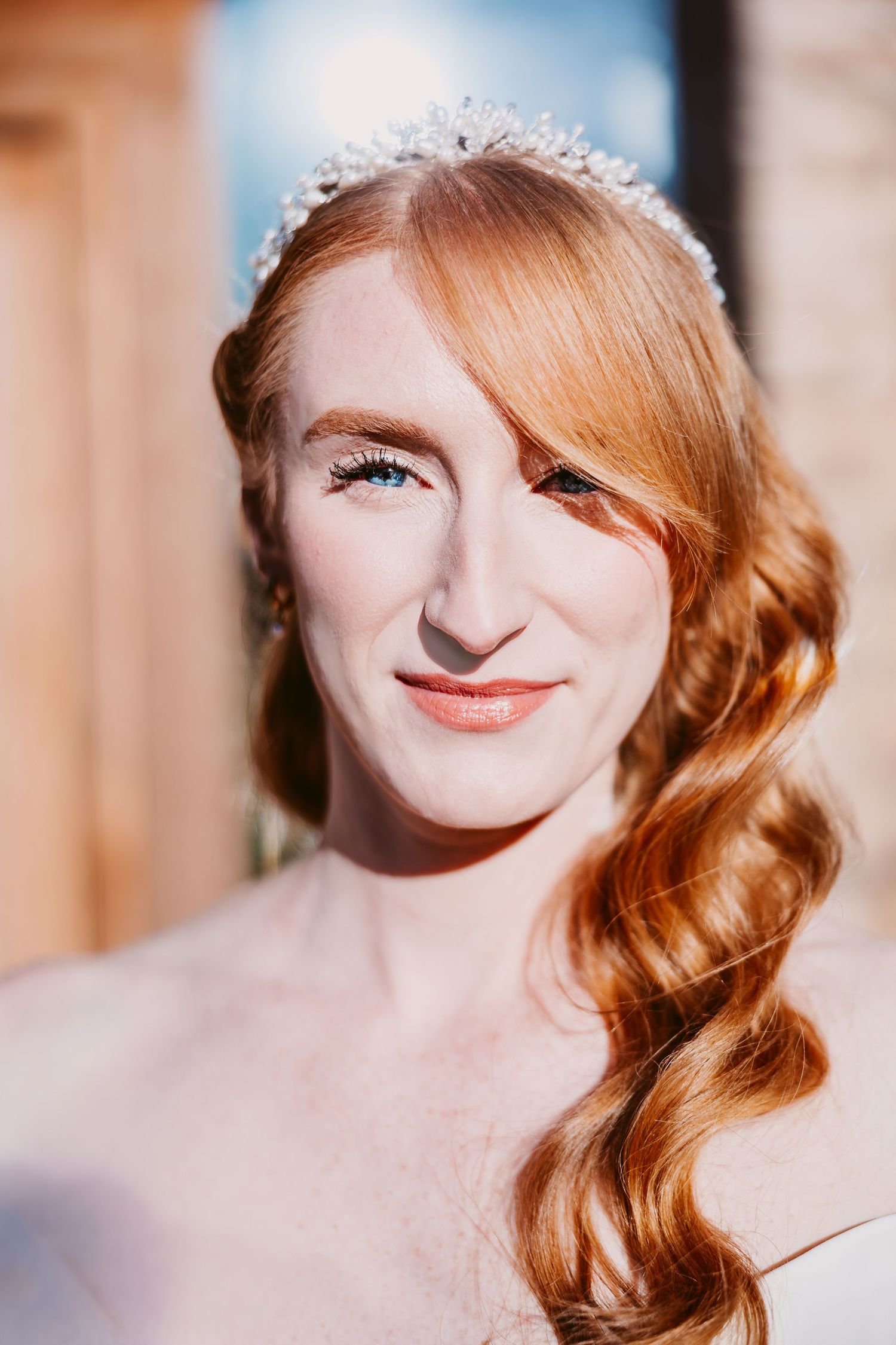 Close-up portrait of a smiling red-haired bride with soft vintage waves and a sparkling crystal tiara in warm sunlight.