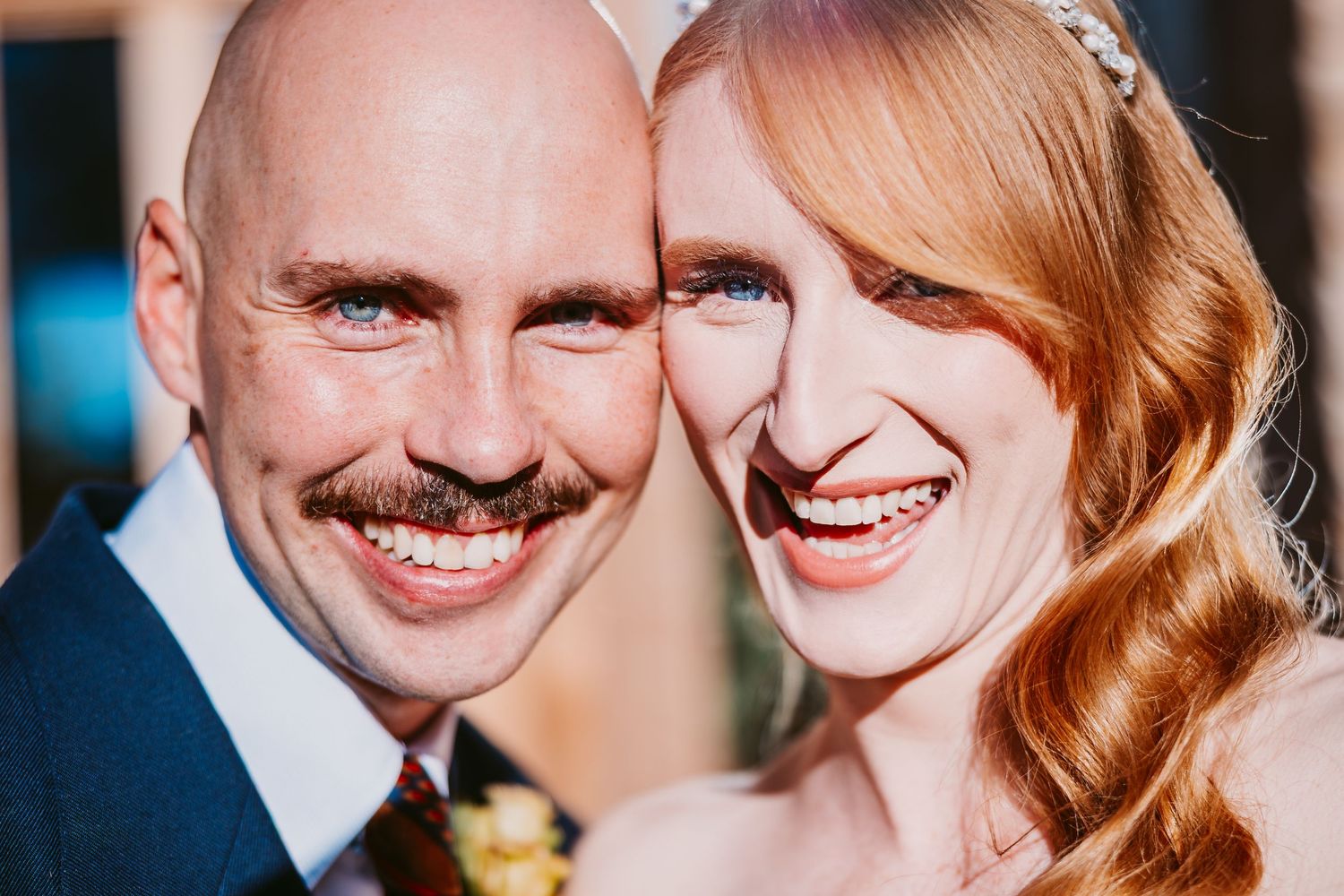 Close-up of a smiling bald groom with a mustache and a red-haired bride with a hair accessory, cheeks pressed together.