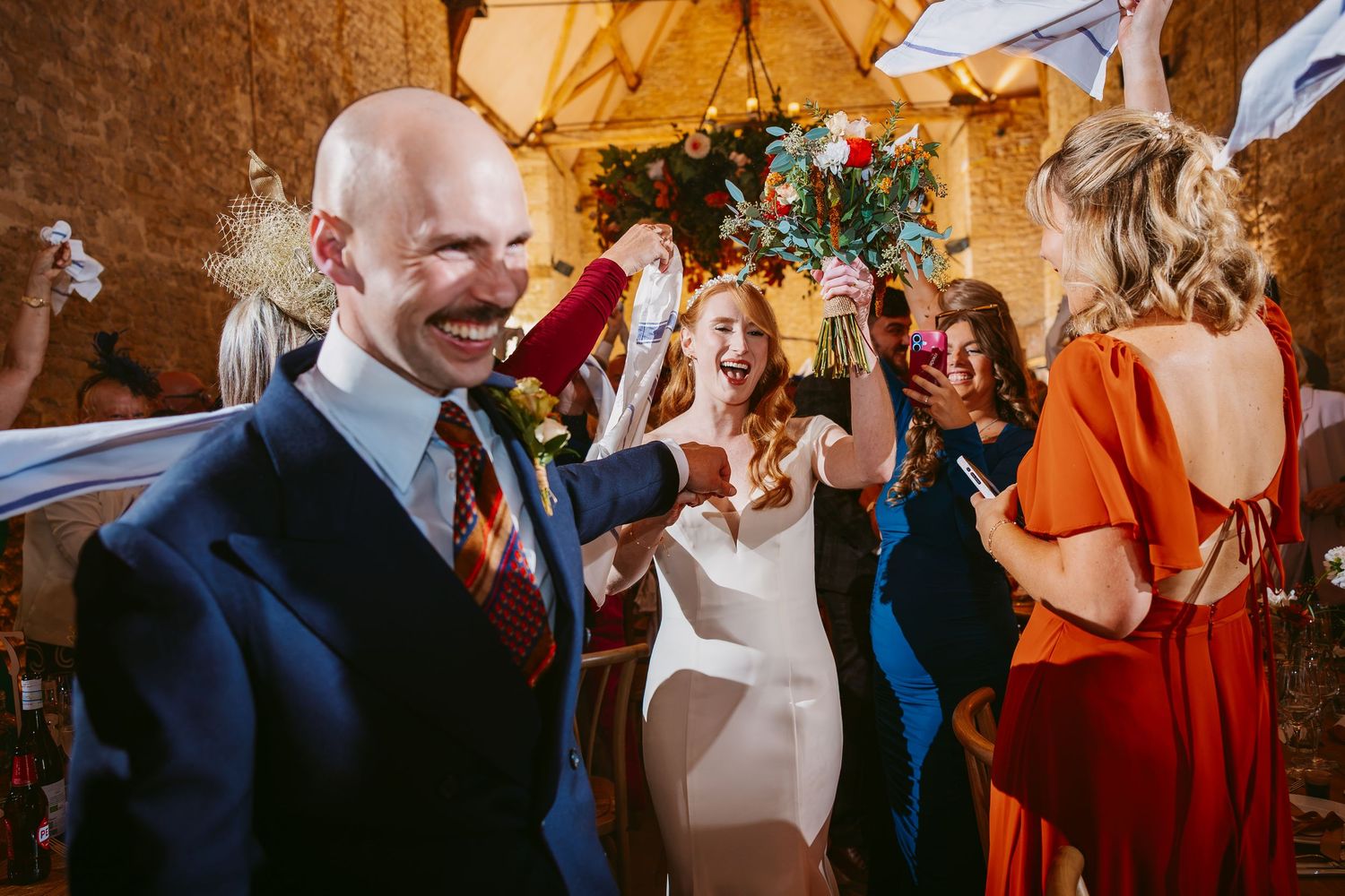 A joyful groom and bride walk through cheering guests holding bouquets and ribbons after their wedding ceremony.