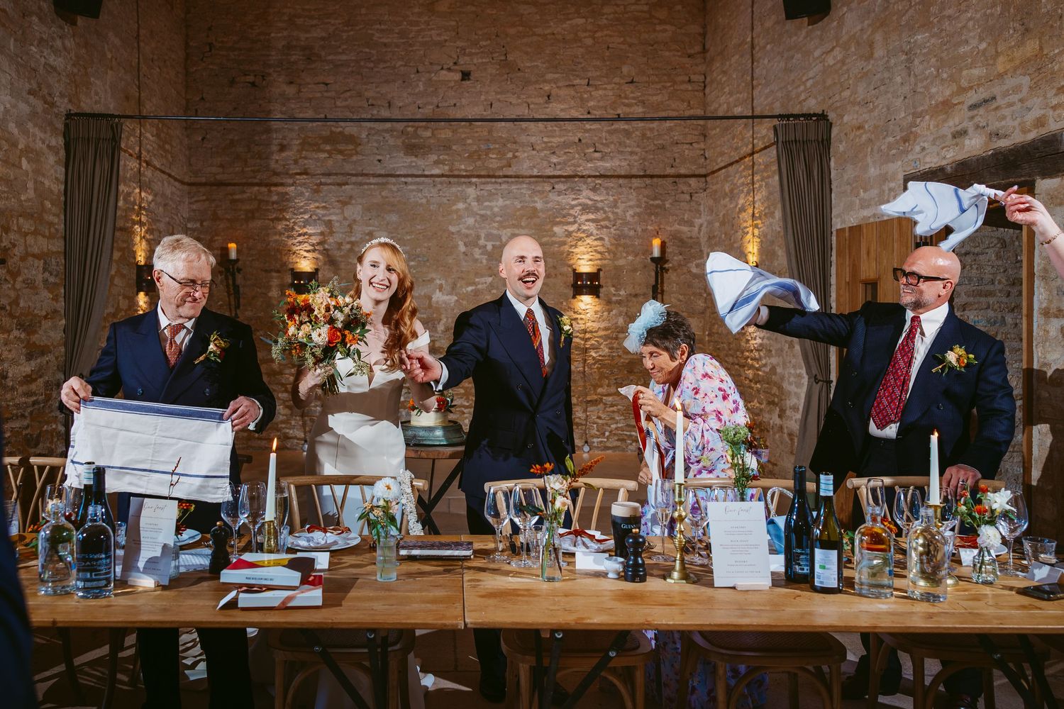 Wedding guests stand at long wooden tables waving white napkins in a candlelit brick barn venue as the bride and groom celebrate.