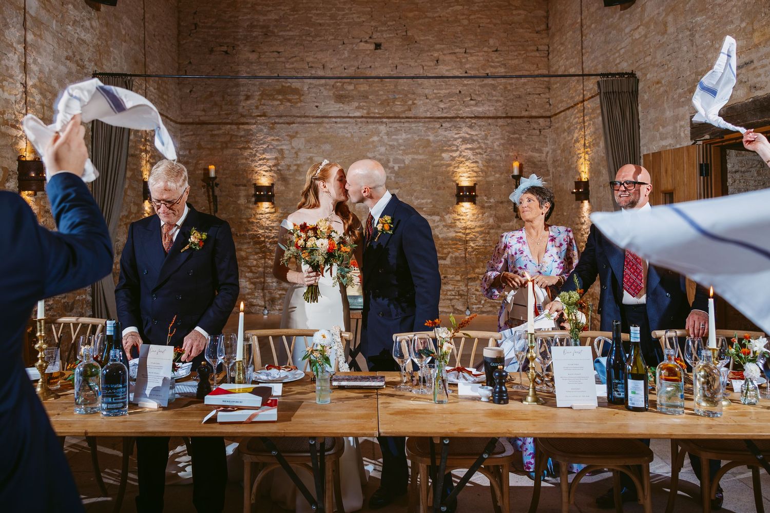 Bride and groom kiss at a reception table as wedding guests wave white napkins in a brick-walled venue with candles.