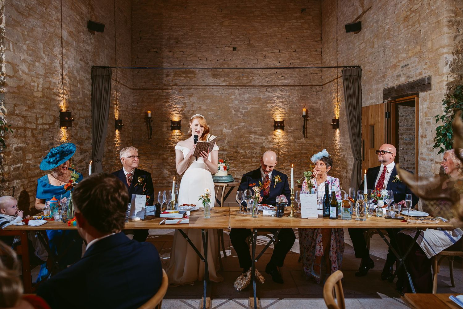 Bride in a white dress stands to give a speech at a rustic stone barn reception while wedding guests watch from long tables.